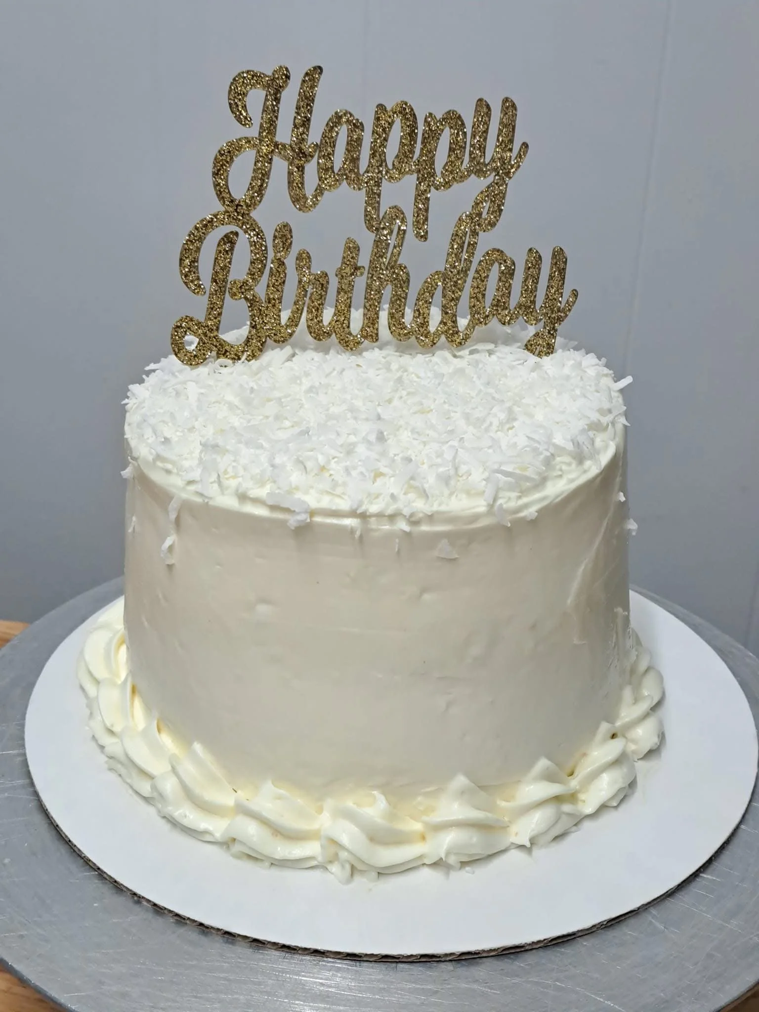 A cake with white frosting, decorated with white chocolate shavings and topped with a gold 'Happy Birthday' cake topper.