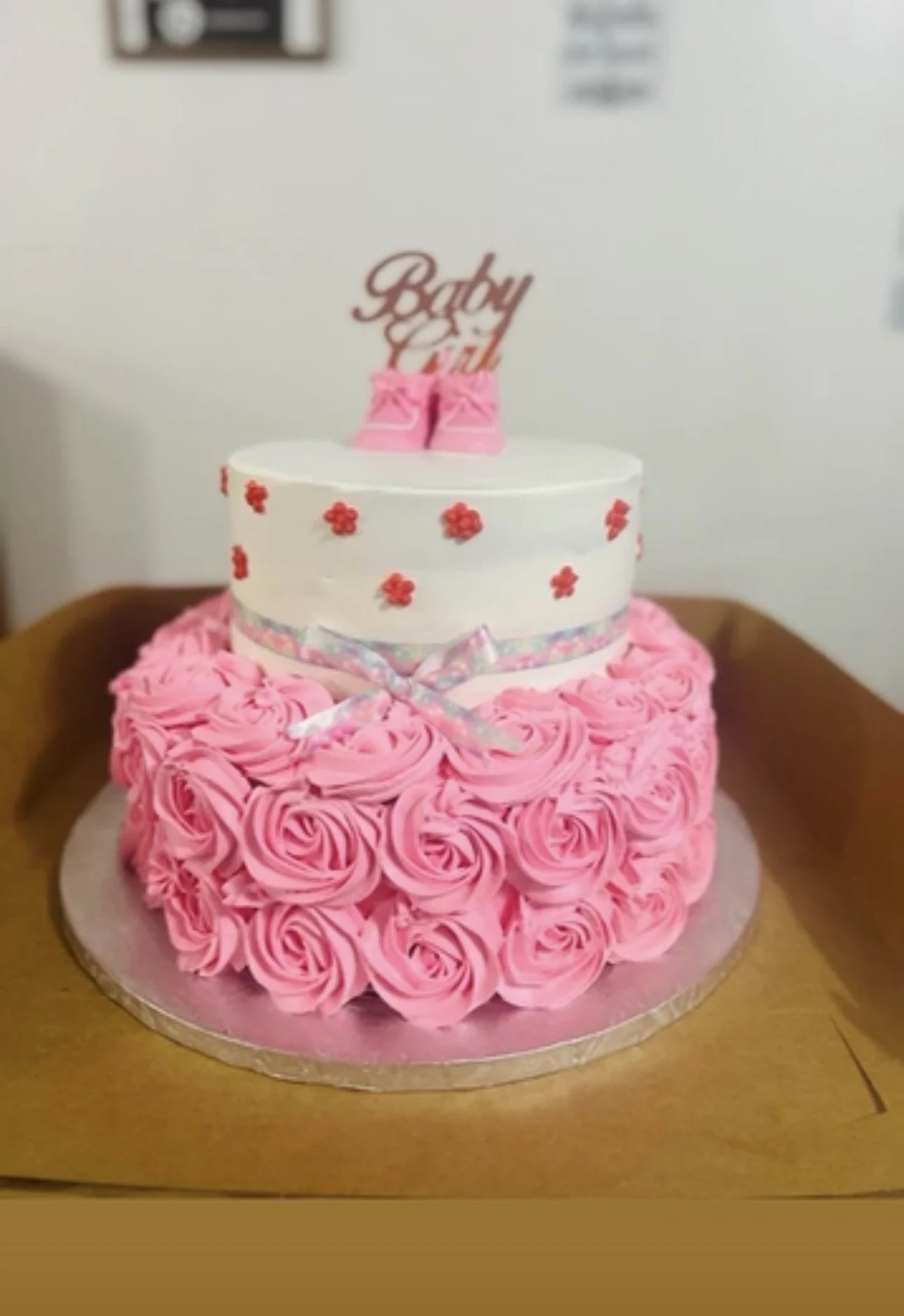 A three-tier pink and white birthday cake with pink frosting roses on the bottom tier, a white middle tier decorated with red flower designs, and a top tier with pink shoes and a topper that reads "Baby Girl."