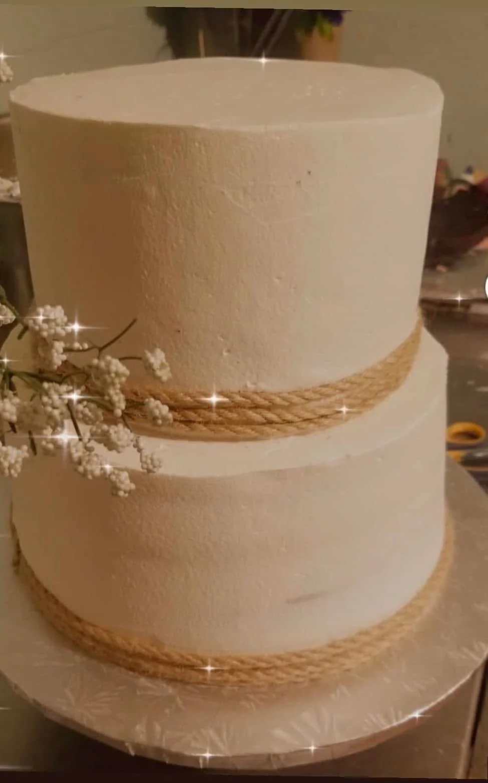 A two-tiered white wedding cake decorated with rope-like borders around each tier, adorned with white flowers on the side, on a round cake board.
