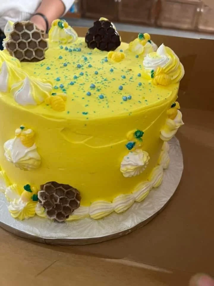 A round yellow cake decorated with honeycomb-shaped chocolate pieces, yellow and white swirled frosting, and sprinkles, sitting on a silver cake board.