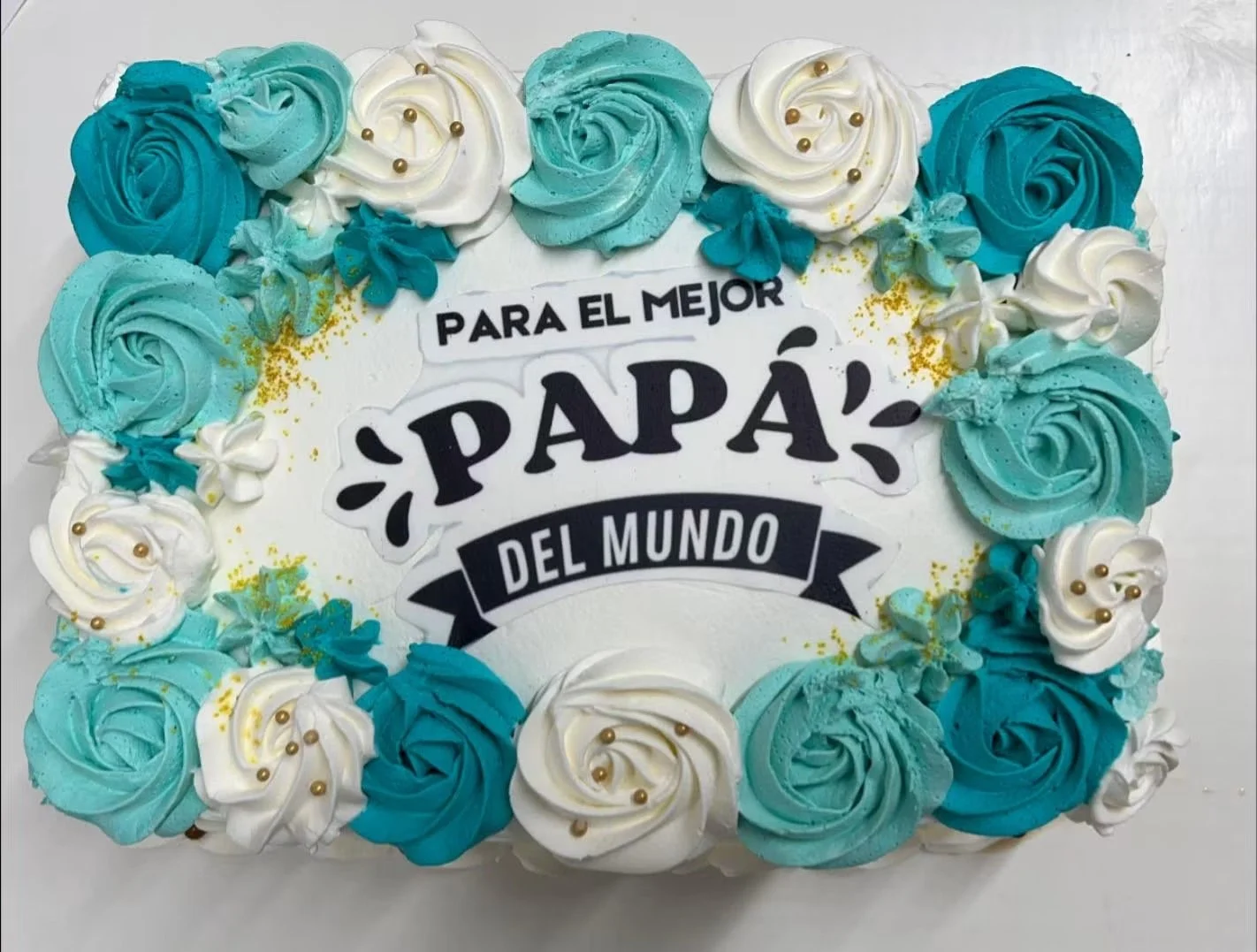 Decorative birthday cake with blue and white swirled frosting roses and gold accents, featuring a message in Spanish that reads 'Para el mejor papá del mundo'.