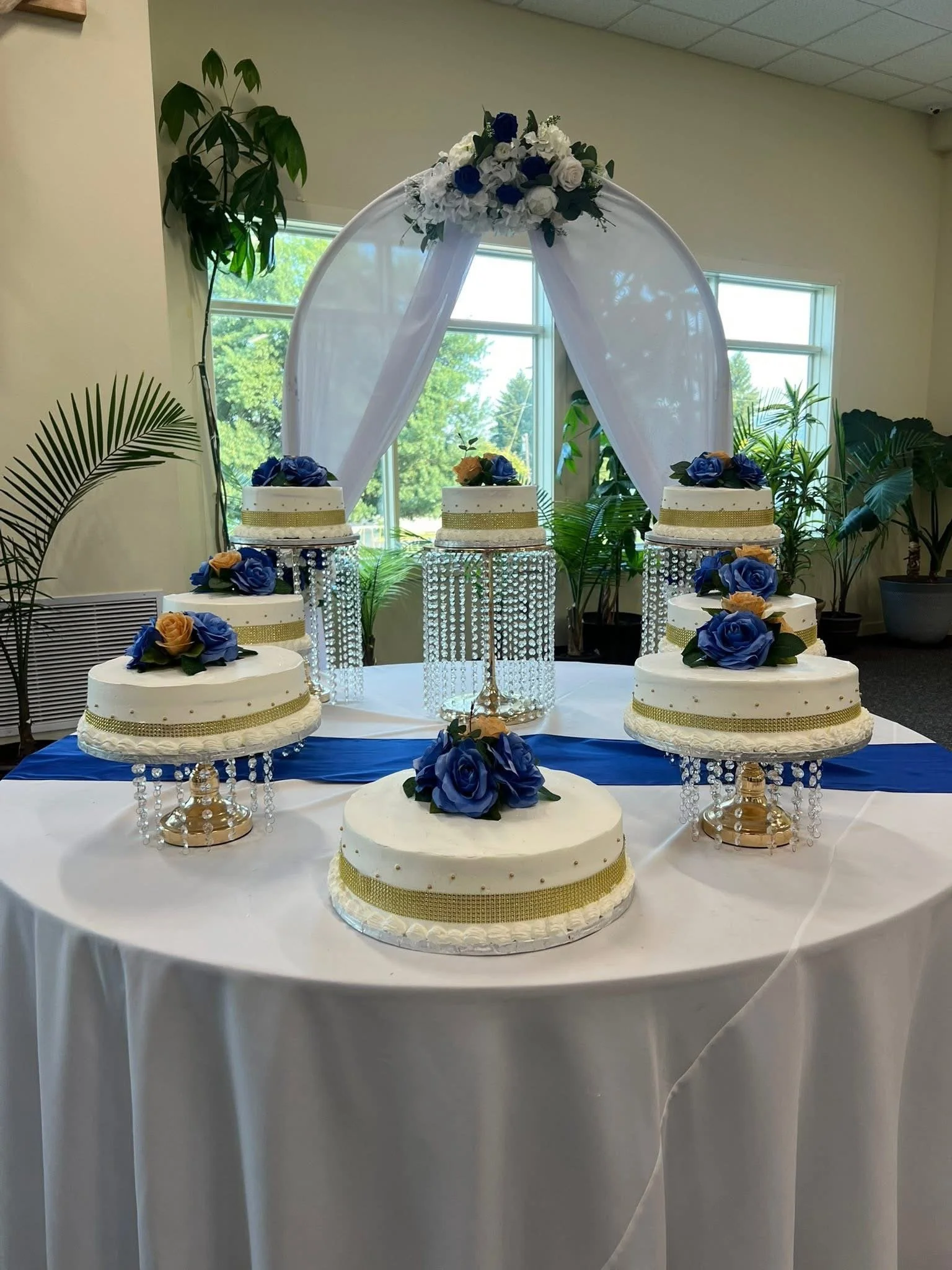 Six white wedding cakes decorated with blue and peach roses, gold ribbon, and hanging crystal beads, arranged on a white and blue tablecloth with a backdrop of green plants and a fabric wedding arch with floral decoration.
