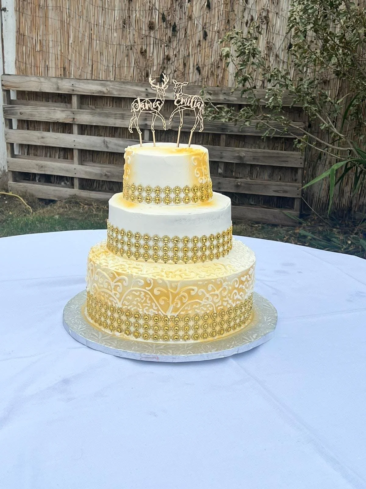 Three-tier wedding cake with gold decorations and intricate lace-like piping, topped with a topper that has two deer with names 'James' and 'Emilia' on their antlers, set on a white tablecloth outdoors.