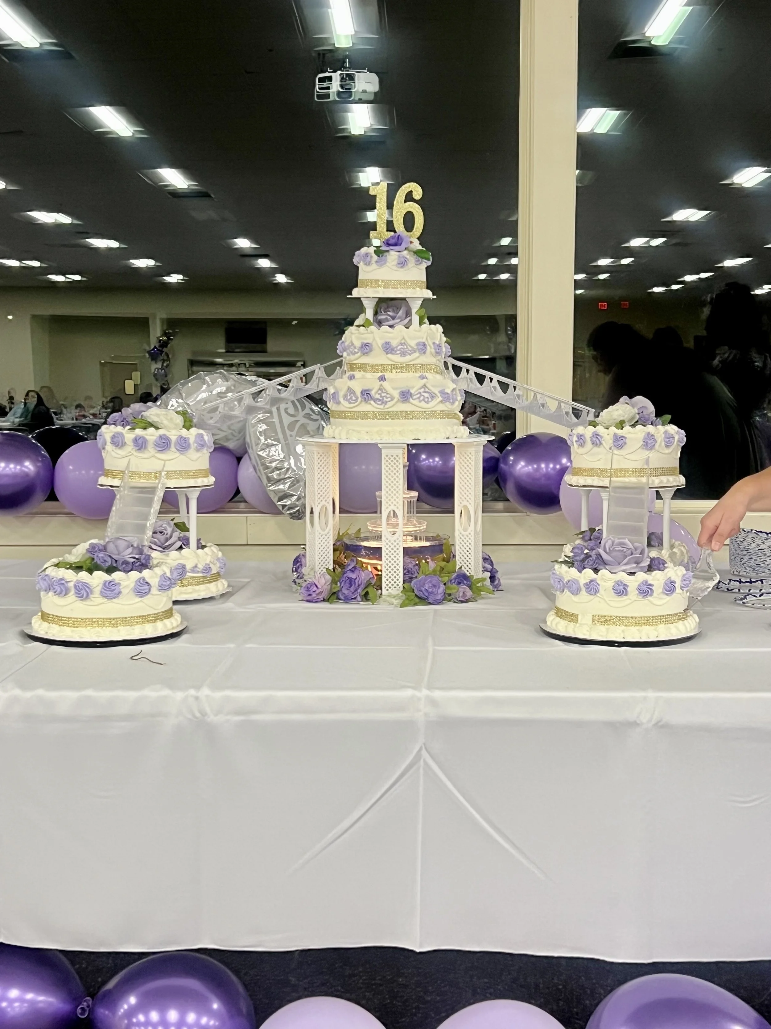 Celebration table with purple and white tiered cake, decorated with purple roses and golden accents, topped with a gold number 16. Surrounded by purple balloons, with a mirror reflecting the cake and balloons, and a few people in the background at an