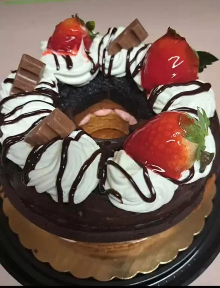 A chocolate cake decorated with white whipped cream, strawberries, and chocolate pieces, drizzled with chocolate syrup.