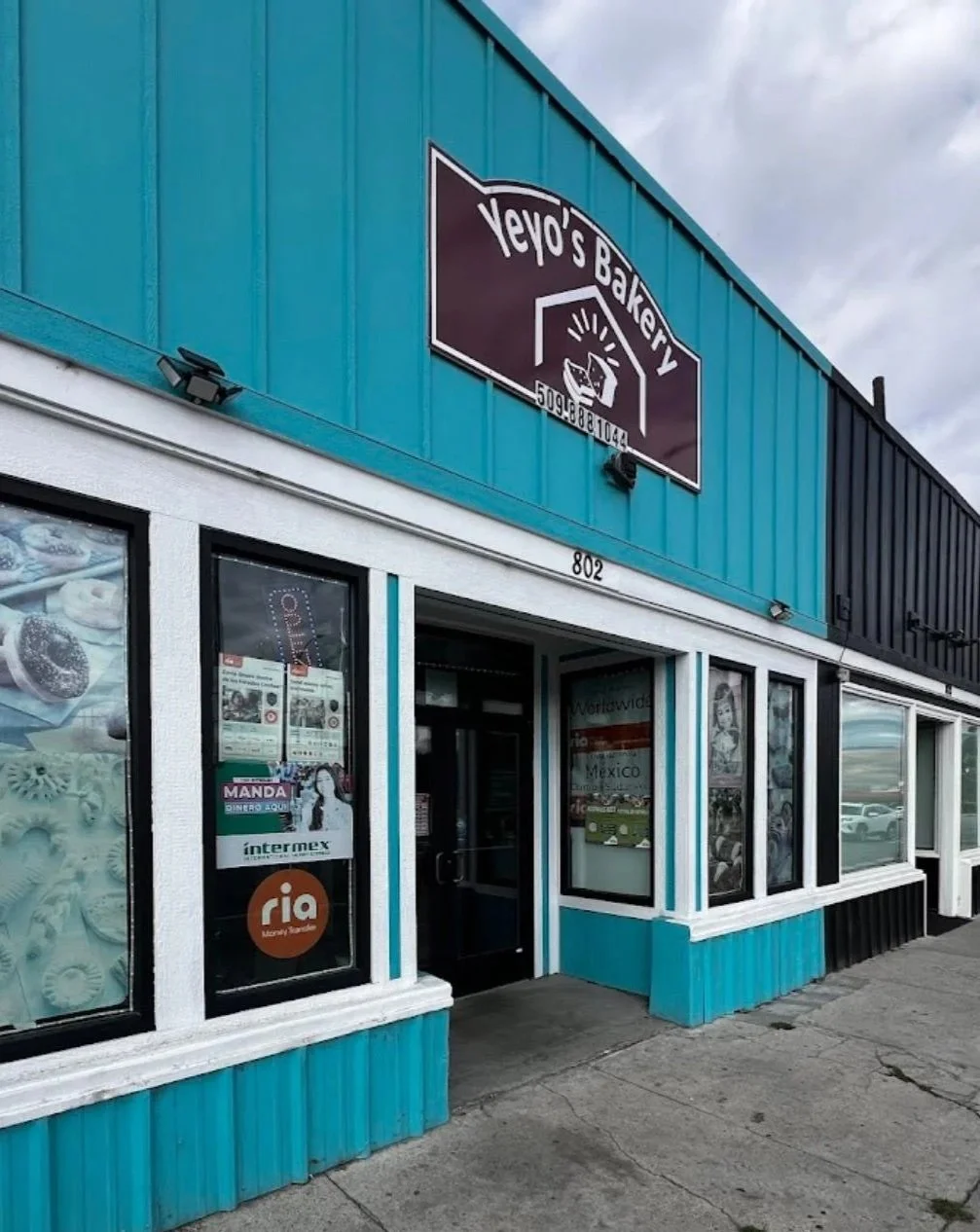 Exterior of a bakery shop with turquoise blue and black siding, a sign that reads 'Yeyo's Bakery,' and large windows with promotional posters.