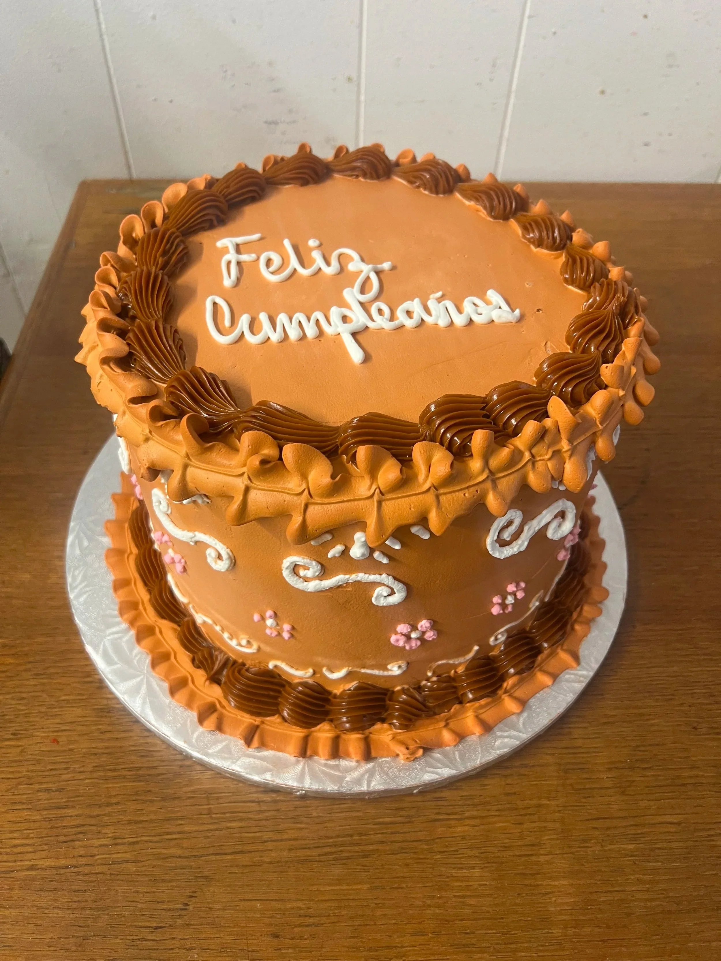 A round cake decorated with orange and brown frosting, with the message "Feliz Cumpleaños" written in white icing on top, placed on a silver tray.