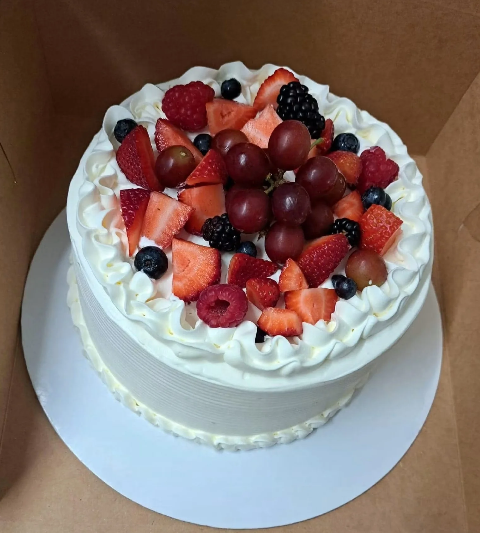 A round white cake decorated with whipped cream and topped with various fresh berries including strawberries, raspberries, blackberries, blueberries, red grapes, and green grapes.