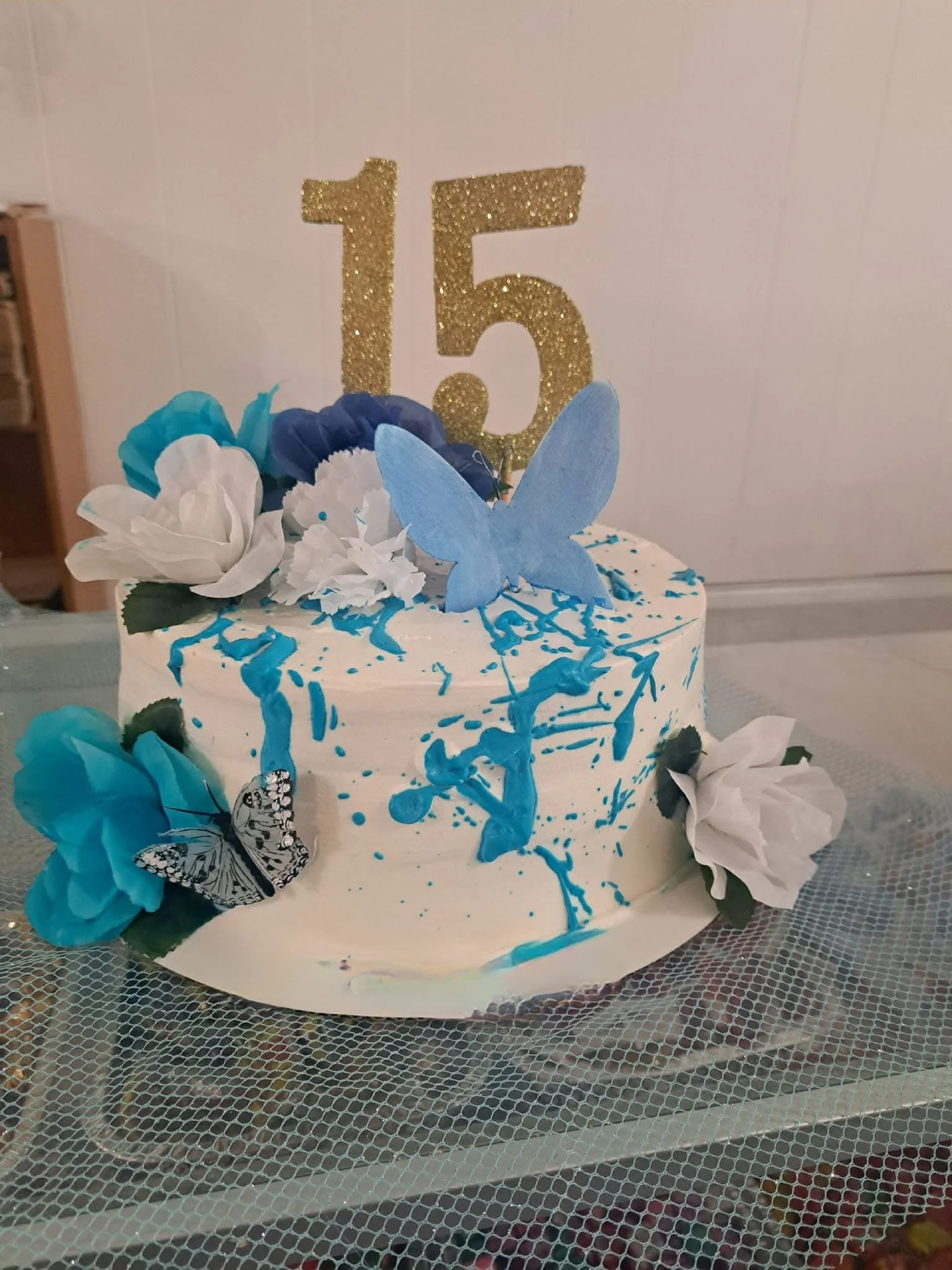Celebration cake with blue and white flowers, a large gold glitter number 15 topper, a blue butterfly decoration, and blue and white sprinkles on top of white frosting.