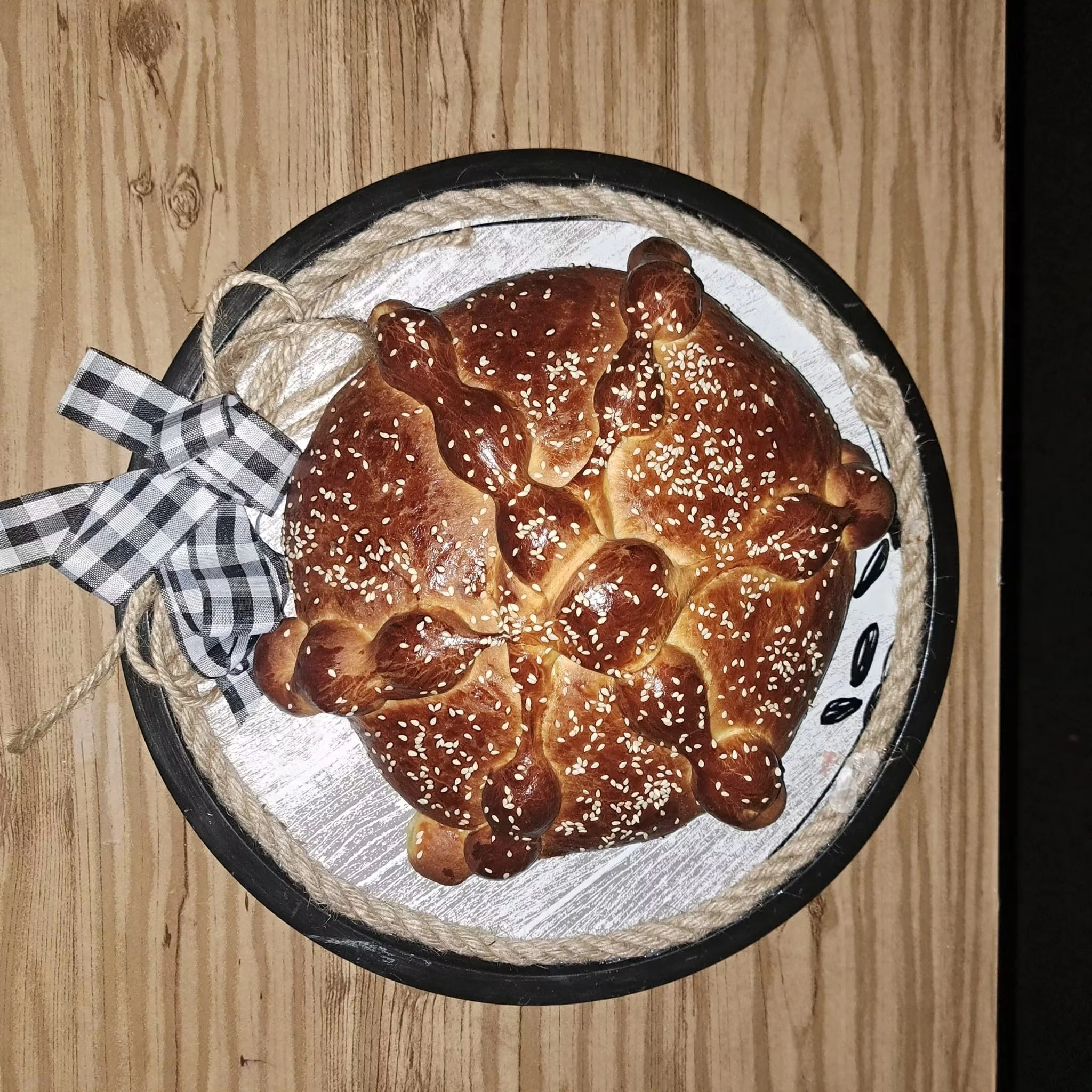 A freshly baked pretzel with sesame seeds on top, placed on a round black plate with a checkered cloth and rope decoration, on a wooden surface.