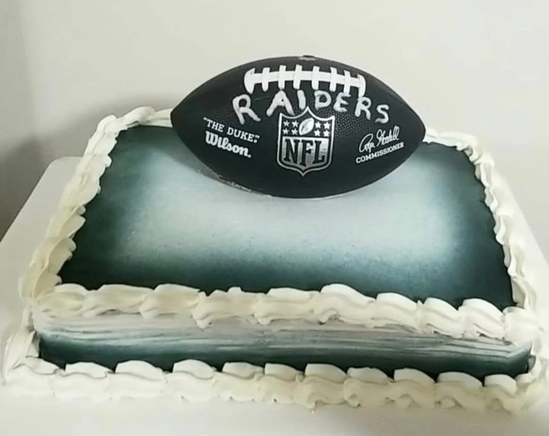 Football-themed cake with a black NFL football on top, decorated with team name Raiders and NFL logo, white frosting border with a blue and gray gradient cake surface.