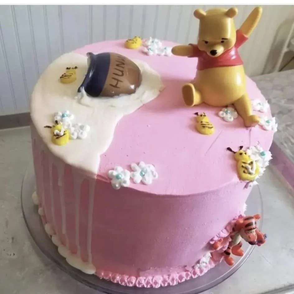 Pink birthday cake with Winnie the Pooh and friends decorations, including a Pooh bear topper, bees, and Tigger, with pink and white icing.