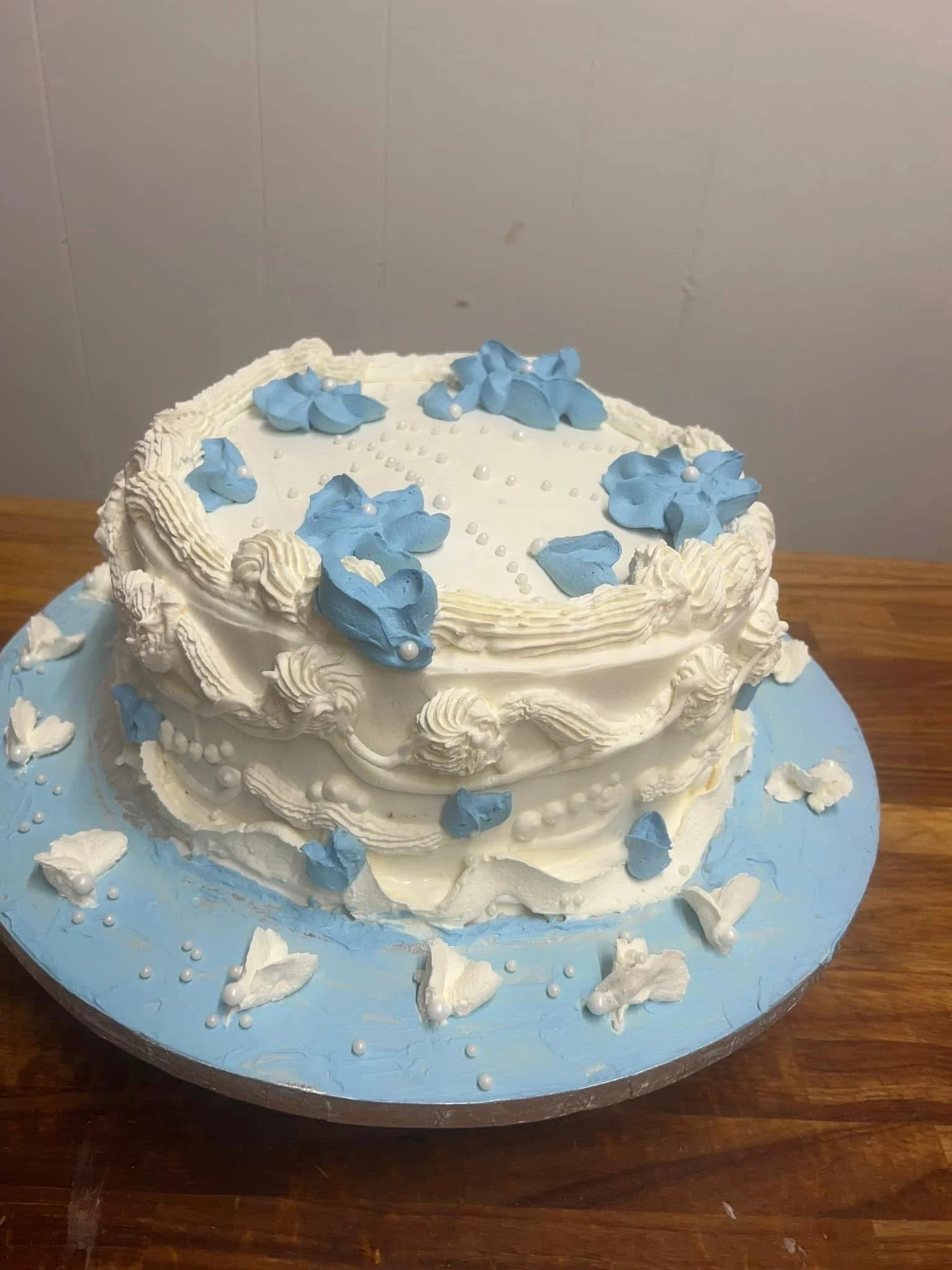 A two-tiered wedding cake decorated with white frosting, blue flower-shaped decorations, and small white pearl-like embellishments. The cake is placed on a light blue cake board with some scattered decoration pieces.