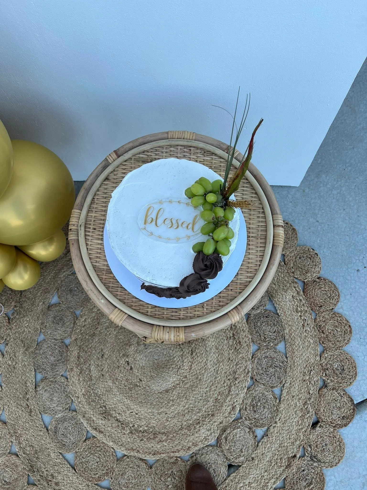 A woven rattan table with a cake decorated with green grapes, brown roses, and a topper that says 'blessed' in gold cursive. A cluster of gold balloons is partially visible on the left, and a woven circular mat is underneath the table. The setting is