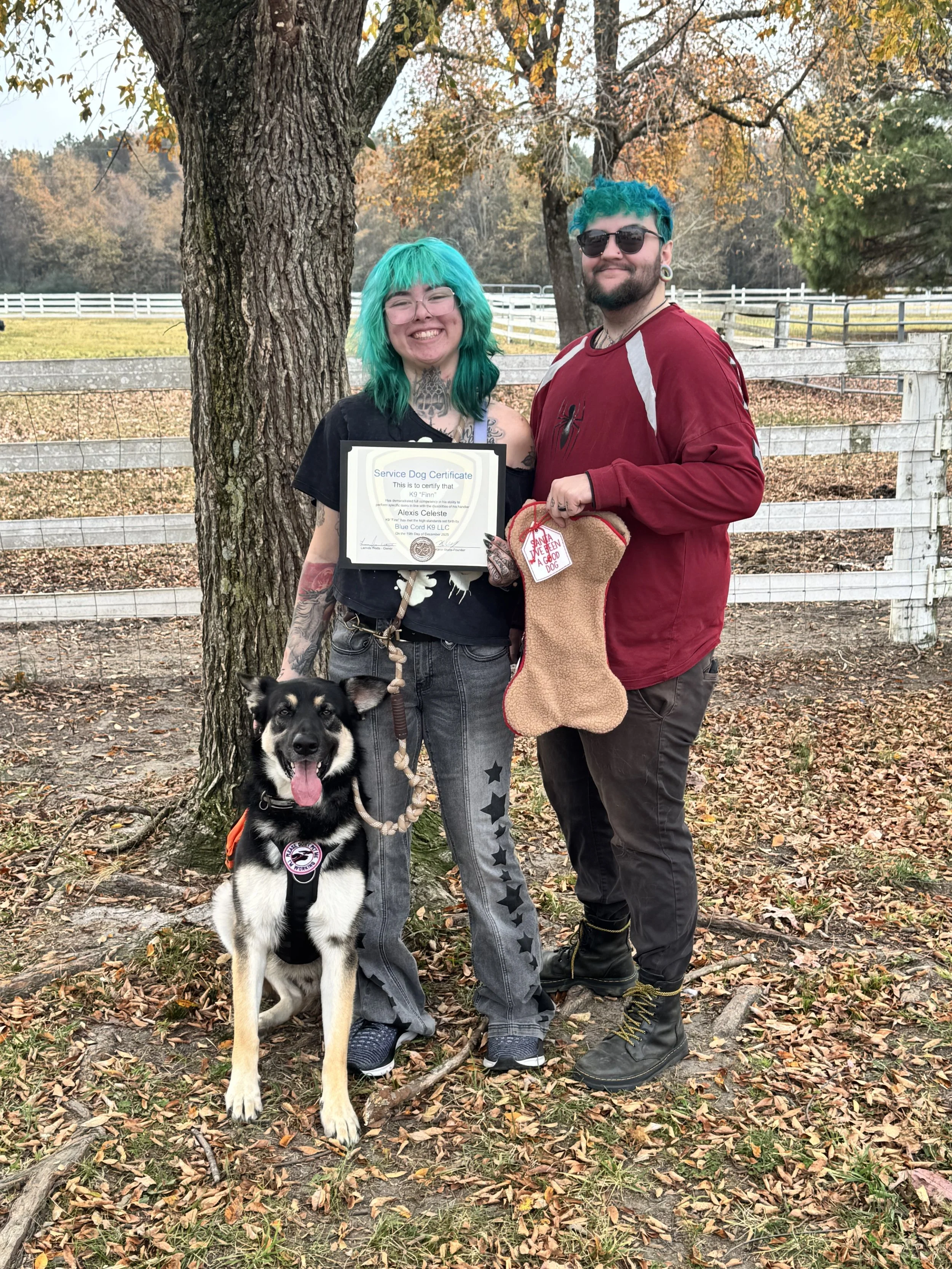A woman with teal hair and tattoos holding a certificate, standing next to a man with teal hair and sunglasses, holding a dog stocking. There is a black and white dog sitting on the ground in front. They are outdoors, under a tree with fallen leaves,