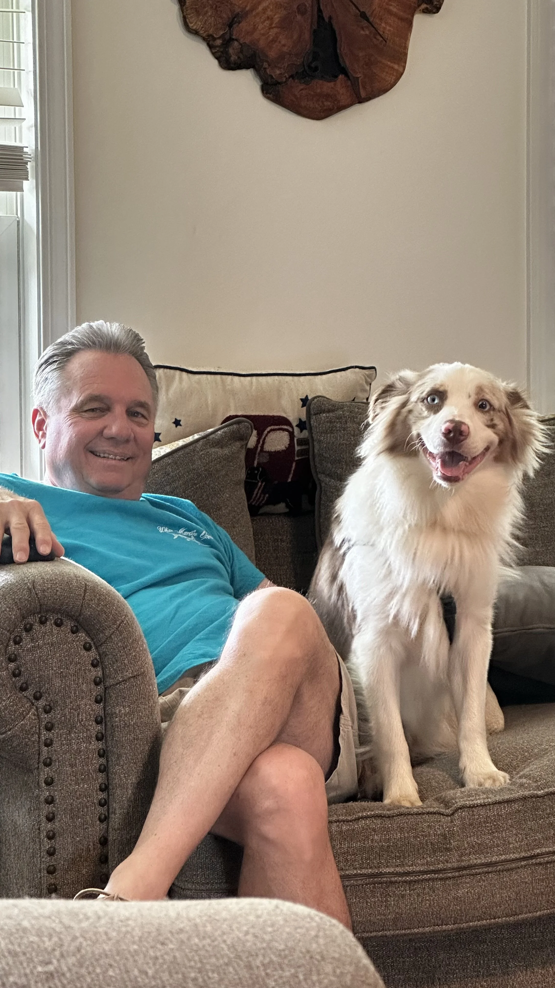 A man with short gray hair smiling while sitting on a brown couch with a Border Collie sitting beside him.