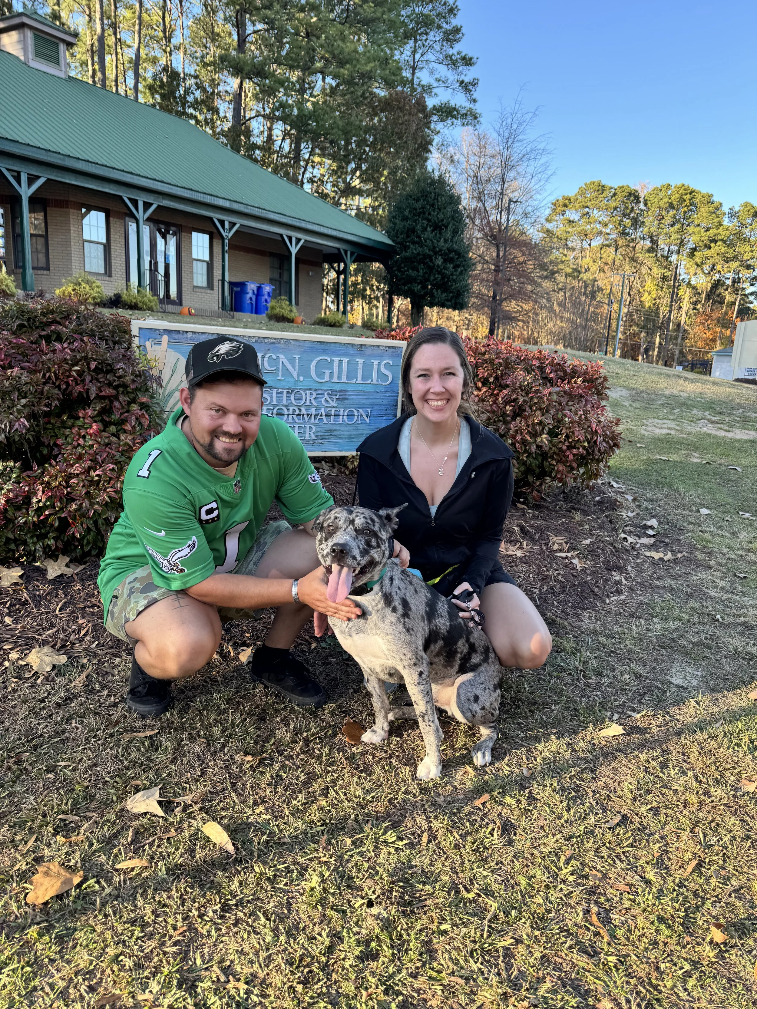 Two people and a dog pose in front of a sign that reads 'Dr. Glicken GILLIS Visitor & Information Center' outside a building with a green roof. The man is wearing a green sports jersey and hat, and the woman is in a black jacket. They are smiling, an