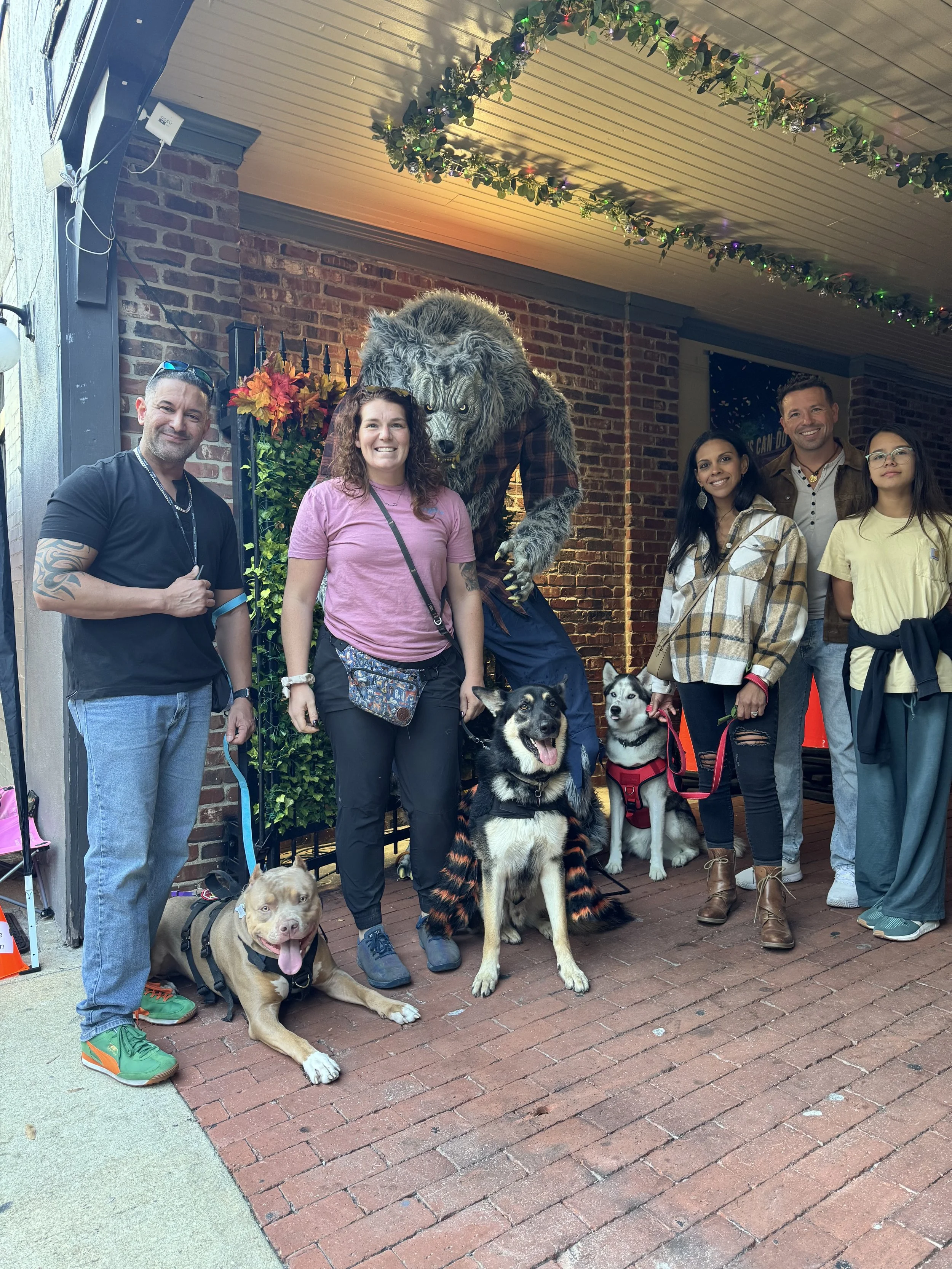 Group of six people and four dogs standing in front of a brick wall, with a wolf mascot costume, some floral decorations, and ceiling garlands overhead.