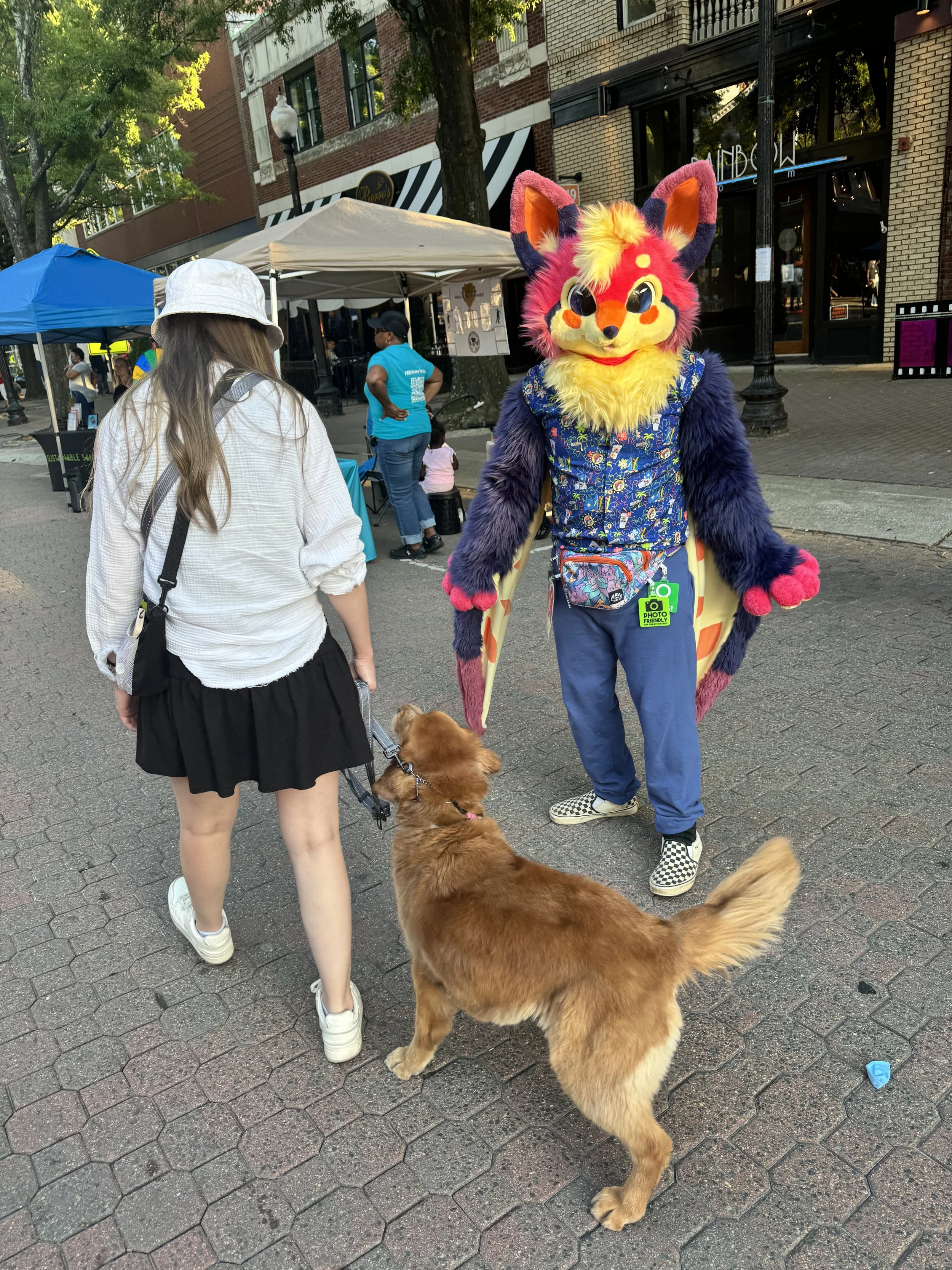 A person in a colorful, furry costume with a large, cartoonish lion or fox head, standing on a city street. They are wearing blue pants, checkered shoes, and a floral vest with a small crossbody pouch. A woman with long hair, dressed in a white shirt