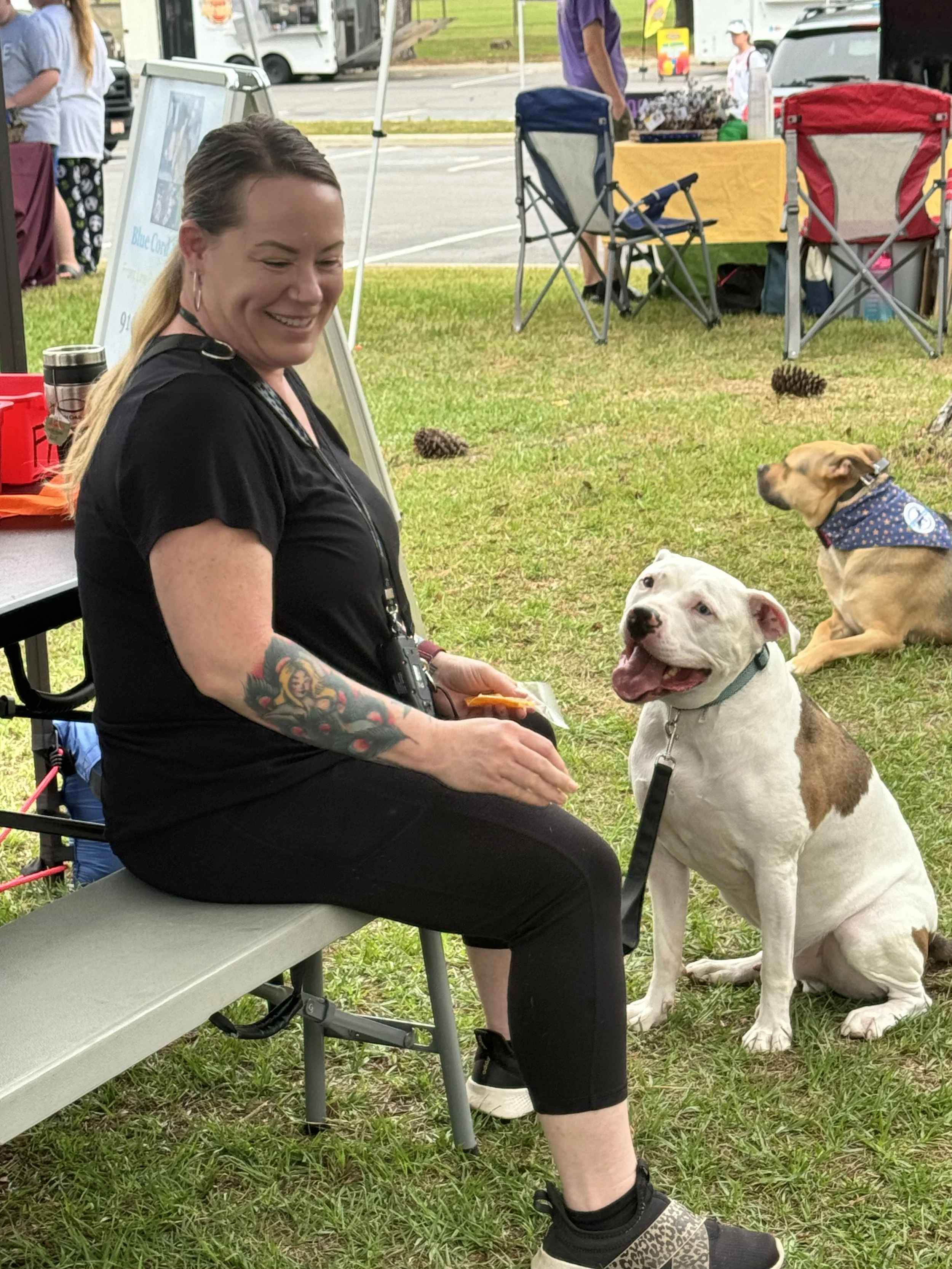 A smiling woman with blonde hair, tattoos on her left arm, sitting on a bench at an outdoor event. She is holding a treat for her white and brown pit bull sitting attentively next to her. In the background, there are tents, chairs, and pinecones on t