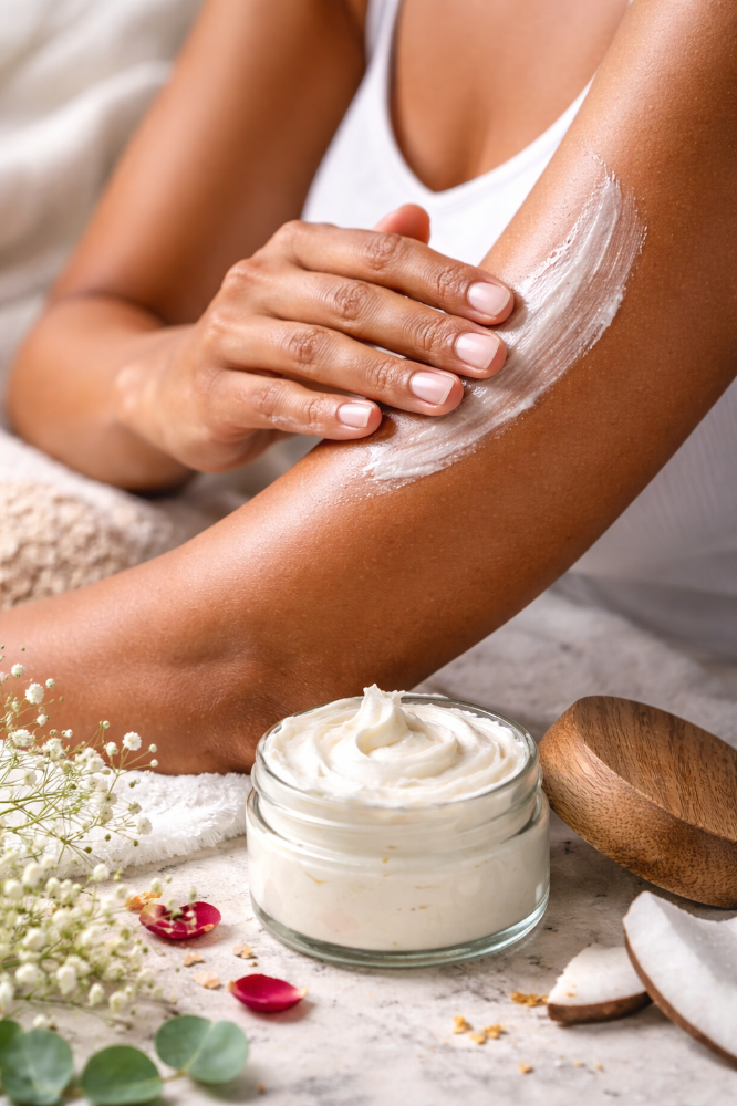 Close-up of a person with a warm caramel complexion gently massaging a rich, whipped body butter into their arm.  The skin appears smooth and radiant as the cream absorbs. In the foreground, an open glass jar of body butter sits on a neutral stone.