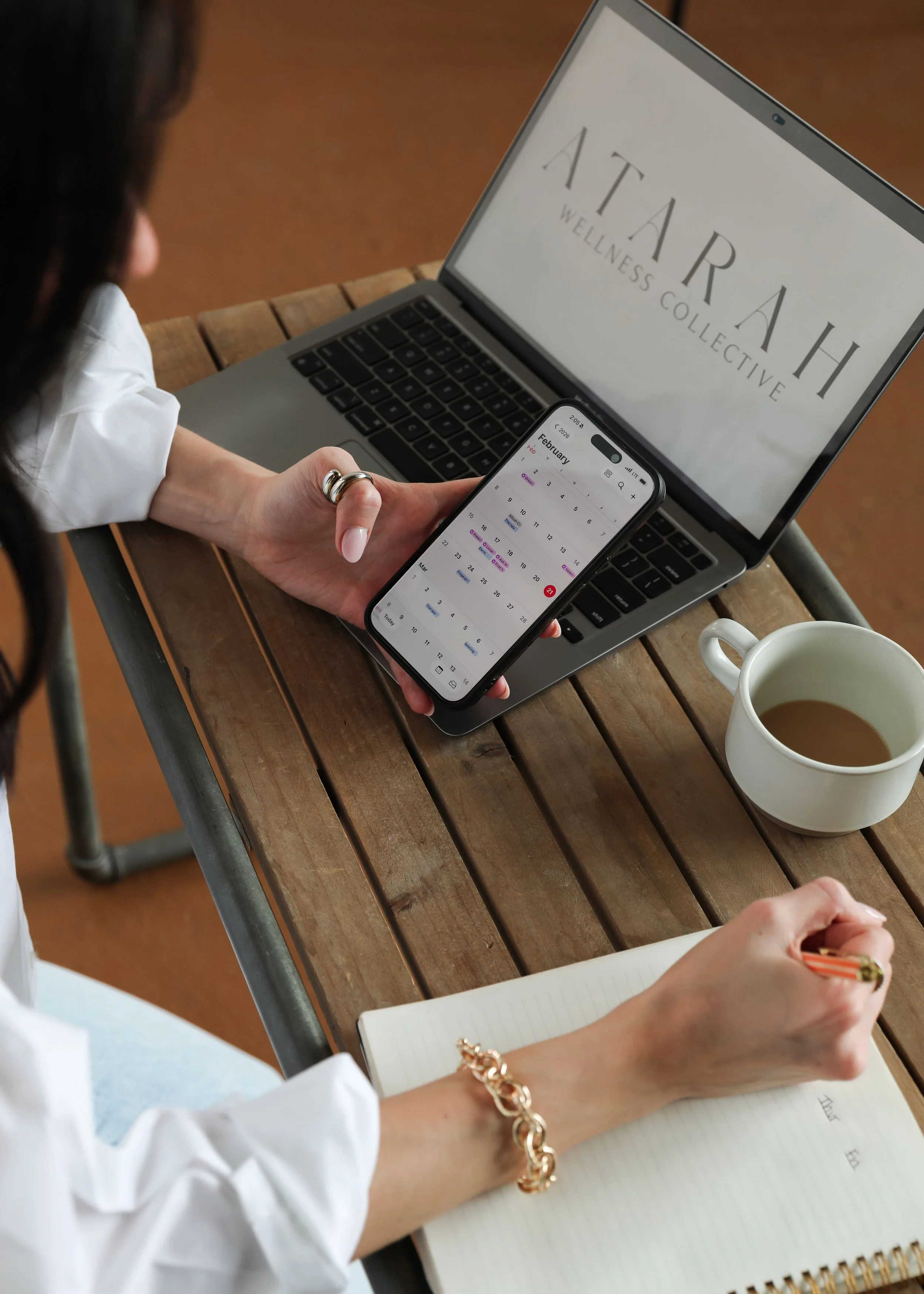 Person sitting at a wooden table with a silver laptop, a smartphone displaying a February calendar, a white mug, and a notebook with a gold pen. The laptop screen shows the words "ATARAH WELLNESS COLLECTIVE."