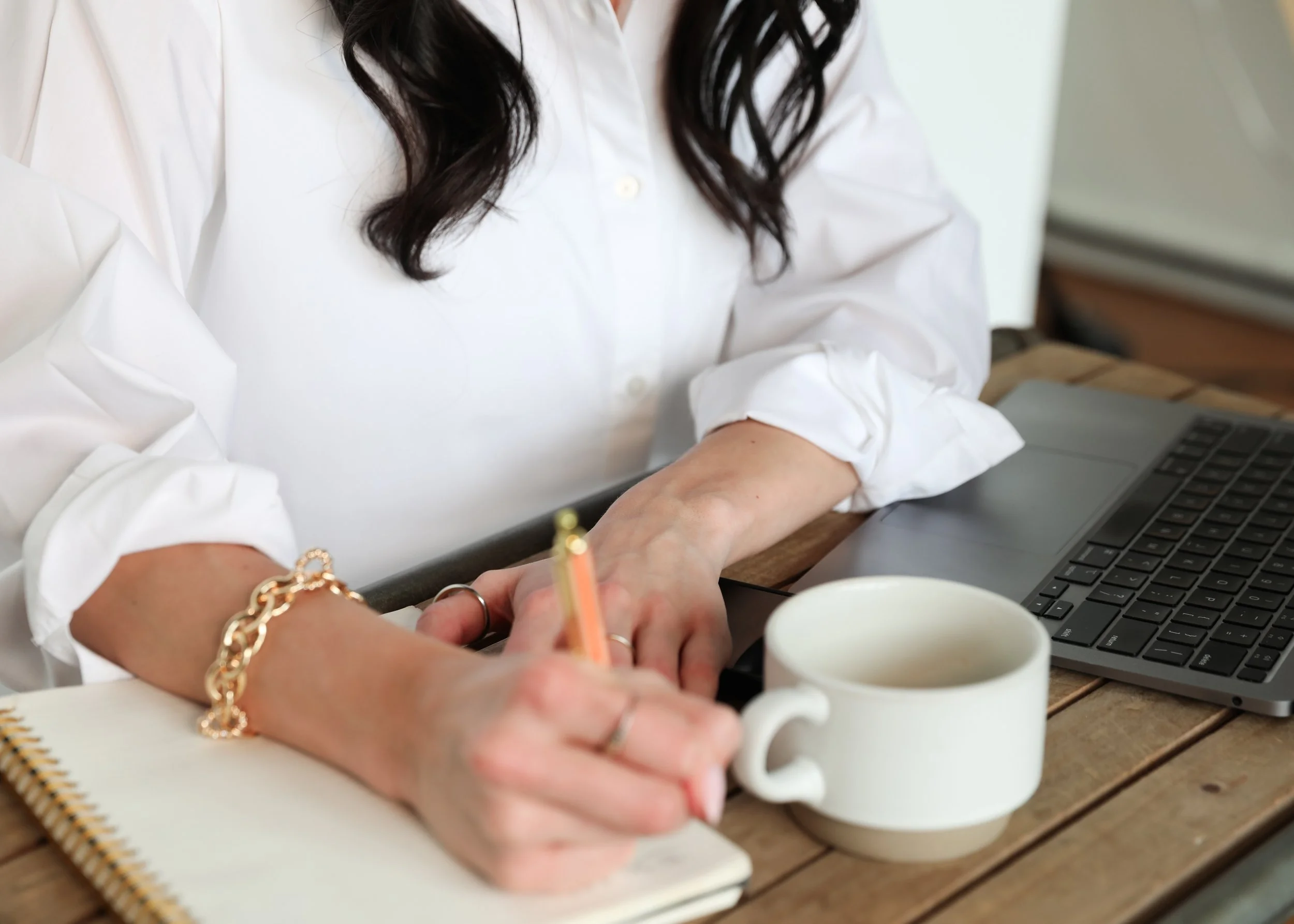 A person dressed in a white shirt with rolled-up sleeves is working at a wooden desk, writing on a notepad with a pencil. There are three white coffee mugs, a laptop, and some other small items on the desk.