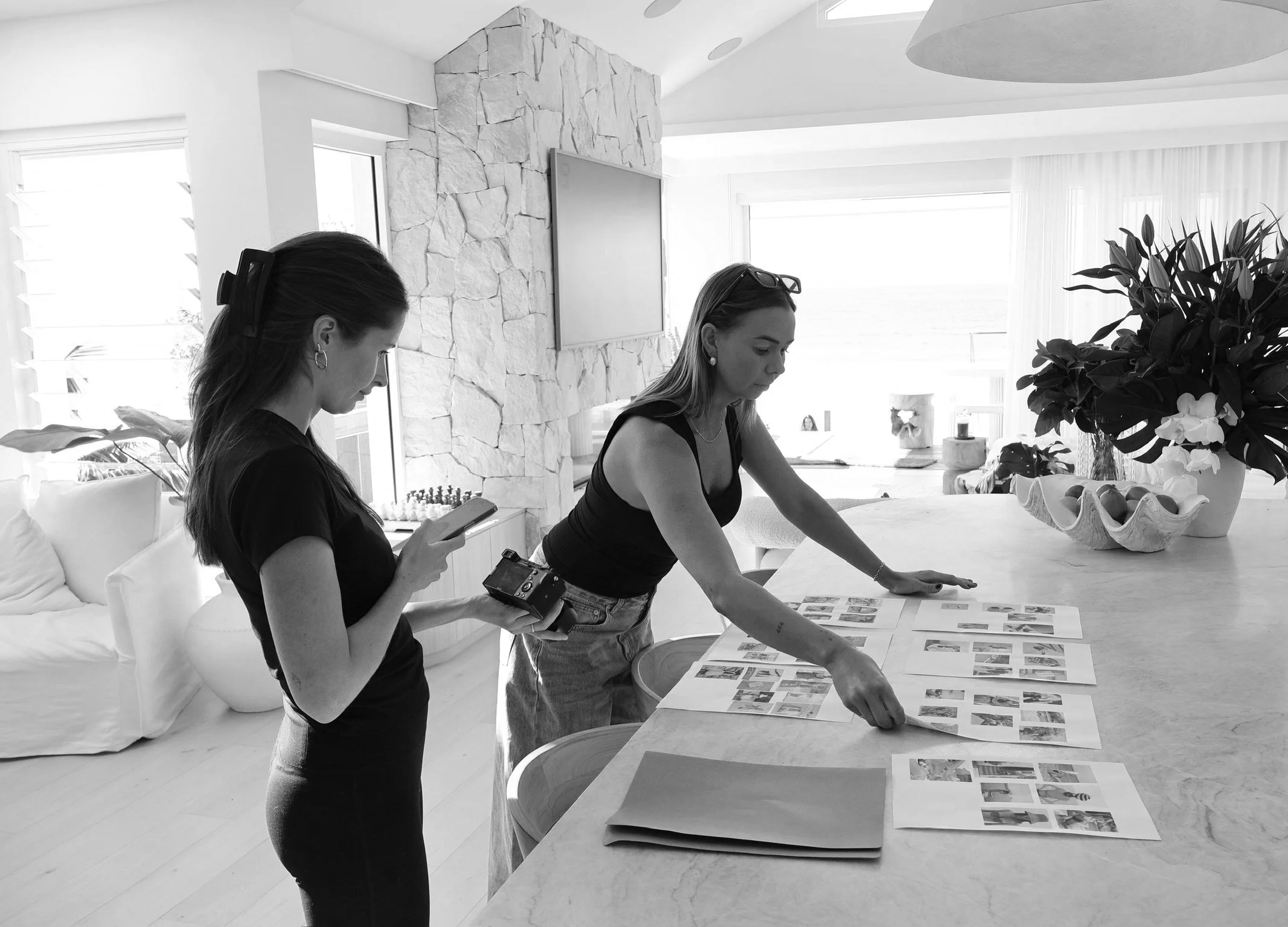 Two women are working at a table covered with printed pages and photographs in a bright, modern room with large windows, a stone accent wall, and indoor plants.
