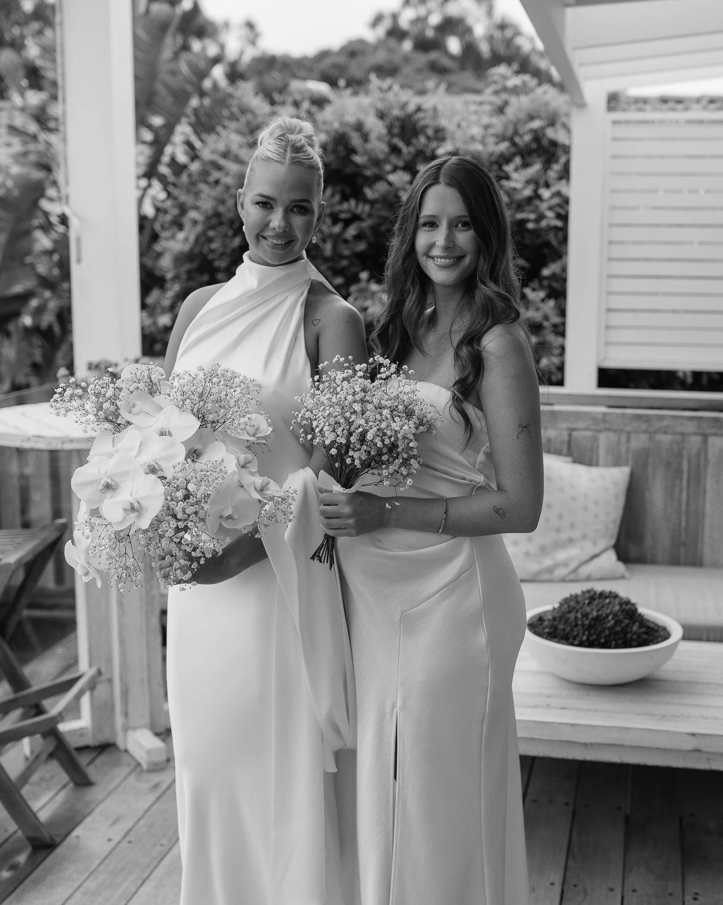 Two women in elegant dresses standing on a wooden porch holding bouquets of flowers, smiling at the camera, with a lush garden and outdoor furniture in the background, in black and white.