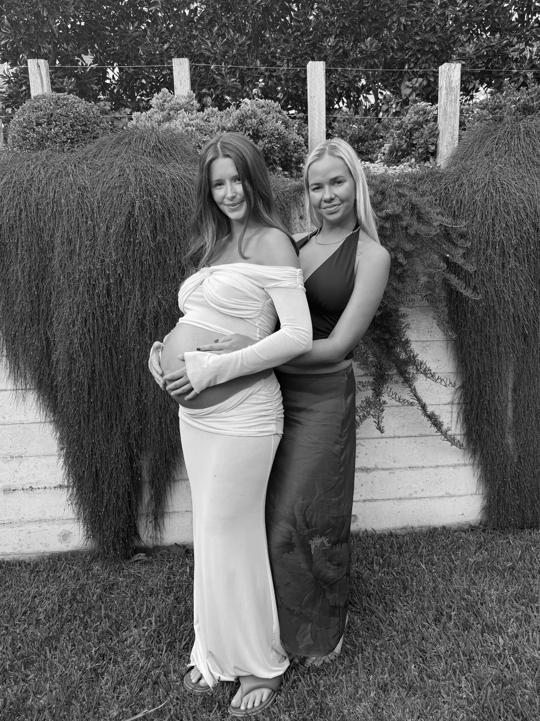 Two women standing outdoors in front of a wooden fence with flowers and bushes, the woman on the left is pregnant and wearing a long, off-the-shoulder dress, the woman on the right has long blonde hair and is wearing a sleeveless dress.