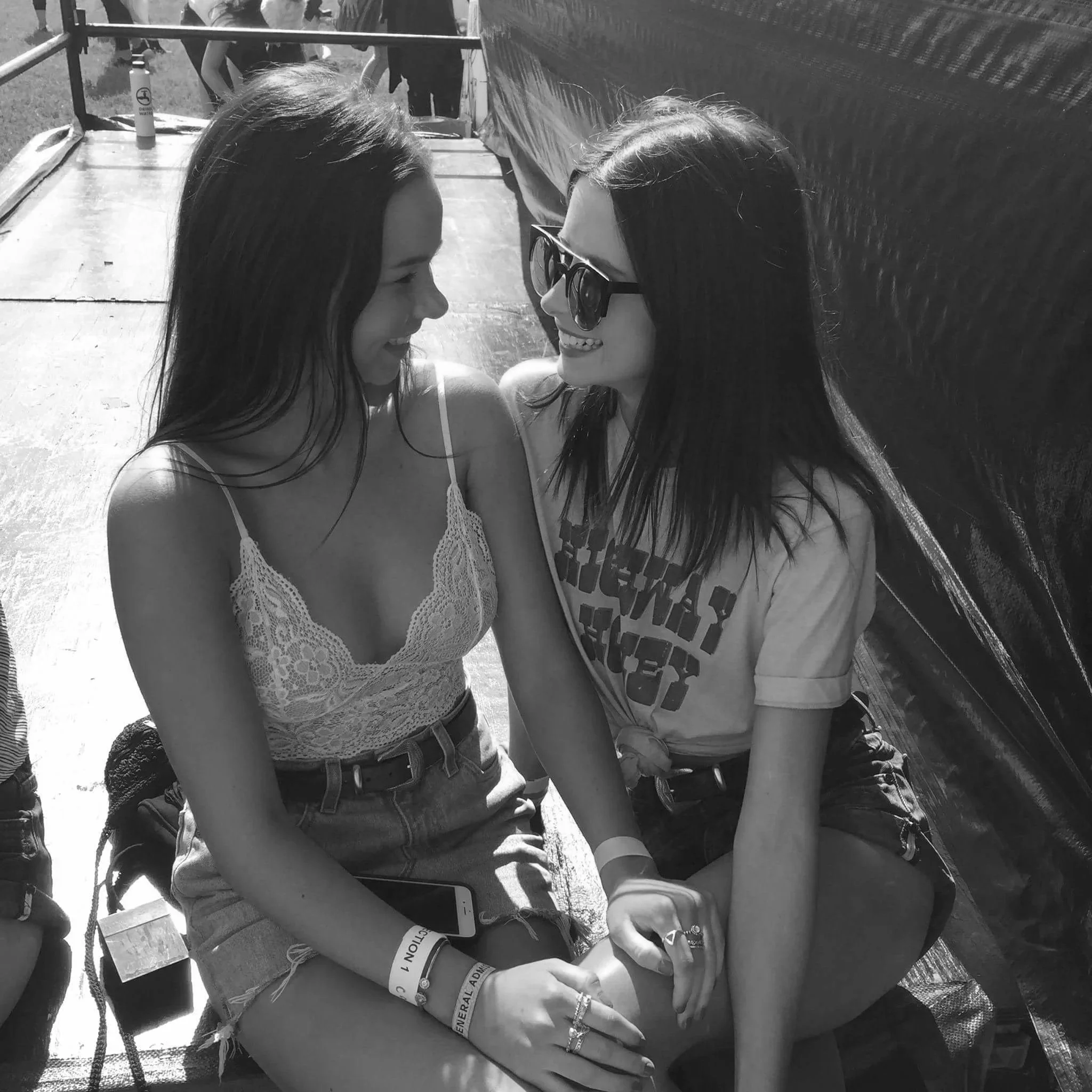 Two young women smiling and leaning their foreheads together on a sunny day. One woman wears sunglasses and a patterned tank top, the other has long dark hair and a bracelet. In the background, there are other people and some equipment, with a focus on their happy interaction.