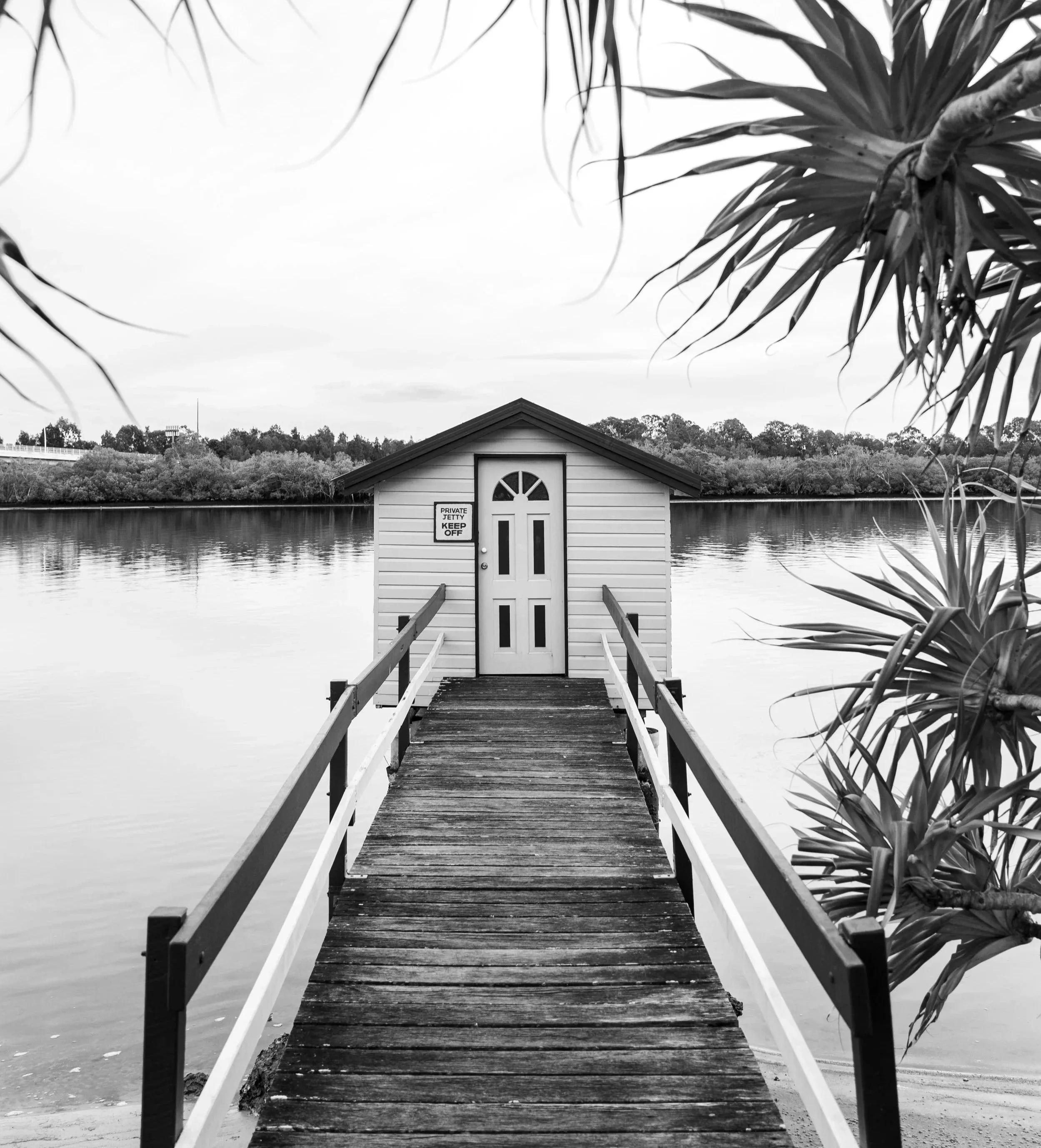 Black and white photo of a small boathouse with a door at the end of a wooden dock extending over a body of water, surrounded by trees and foliage.