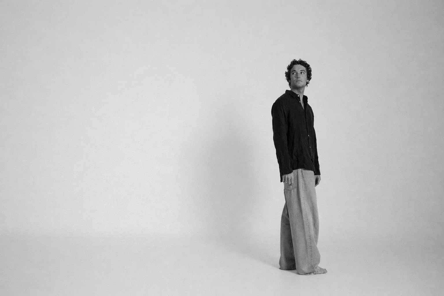 A young man with curly hair wearing a dark shirt and light jeans standing in a vast, empty white space.