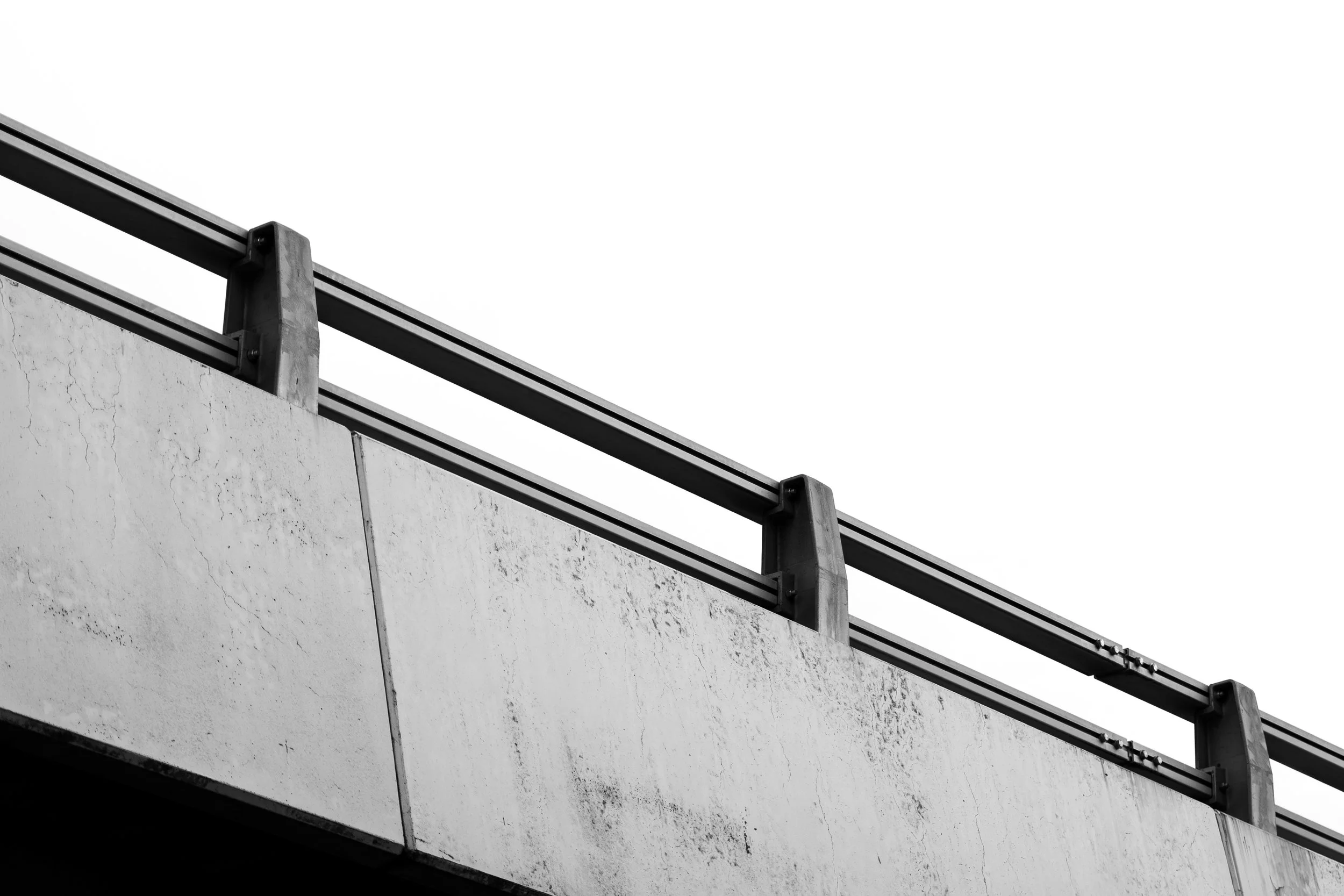 Close-up black and white photo of a concrete bridge railing with metal supports, showing weathered surface and textured concrete, against a clear sky.