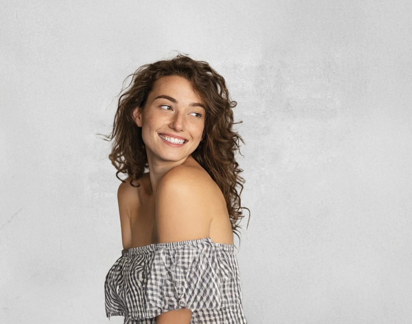 A young woman with curly brown hair smiling and looking to her left, wearing an off-shoulder checkered top, against a plain light gray background.