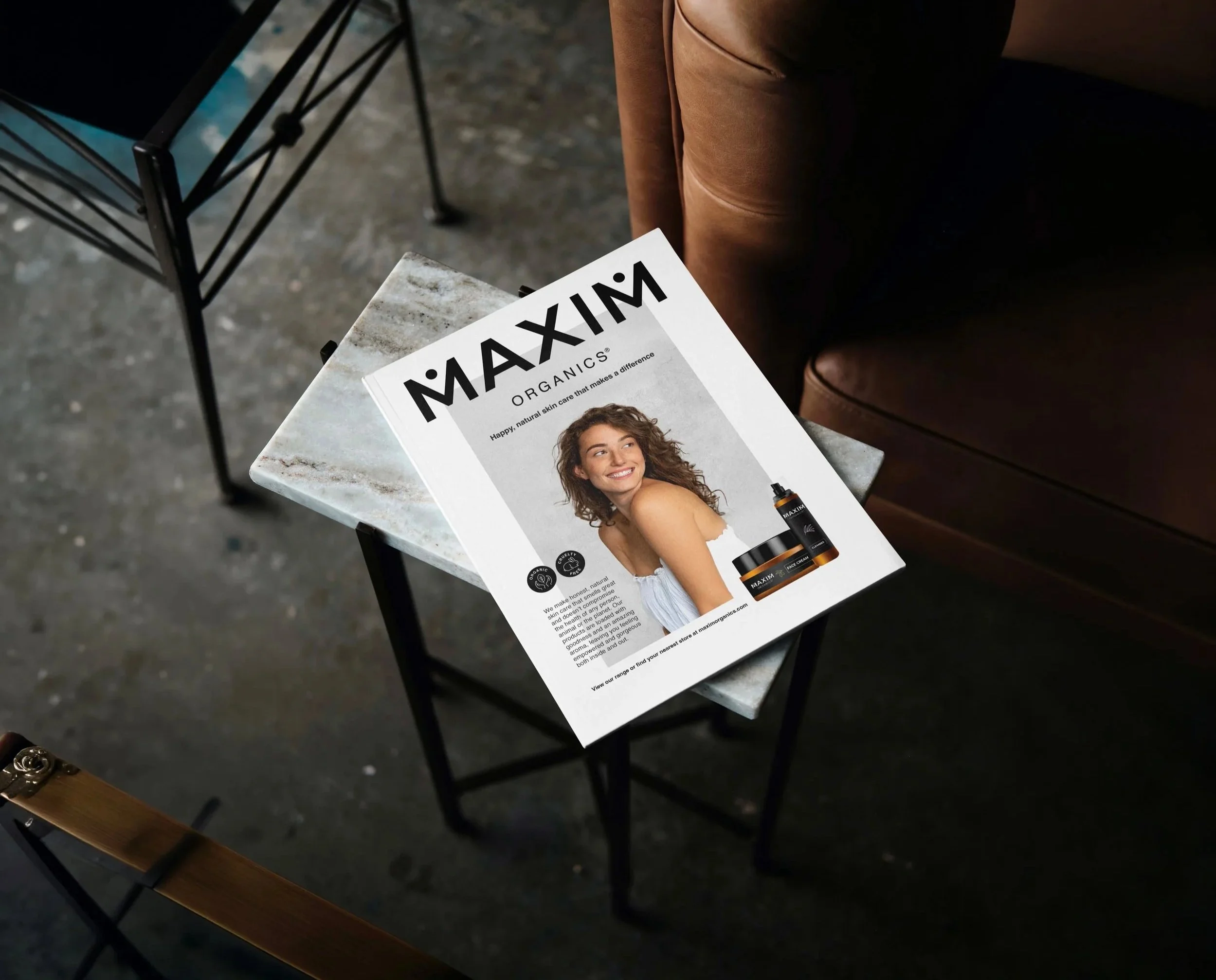 A magazine titled 'MAXIM ORGANICS' on a small marble-top table next to a brown leather armchair. The magazine features a smiling woman with curly hair and pictures of skincare products.