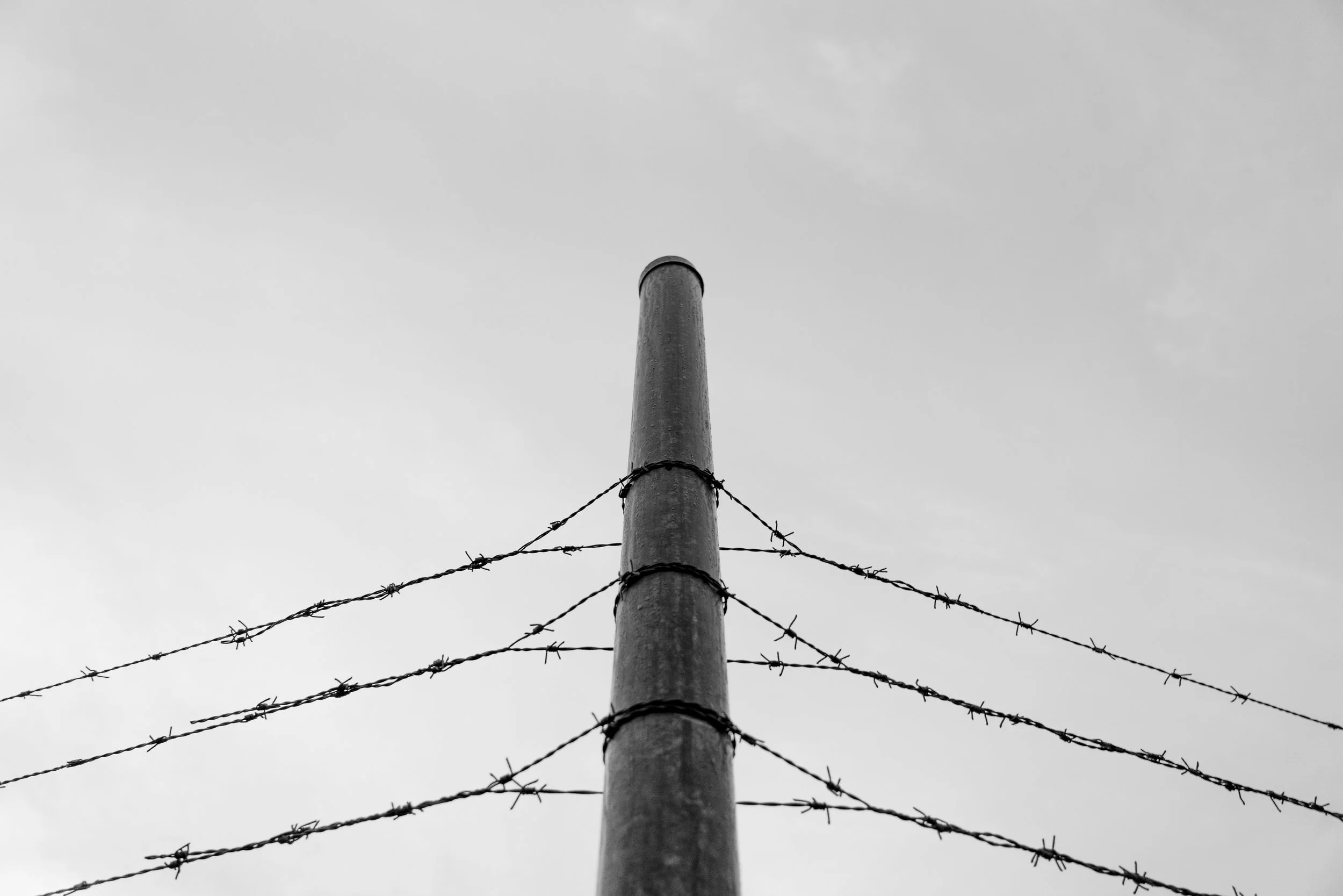 A tall wooden utility pole with barbed wire fencing, seen from below, against a cloudy sky.