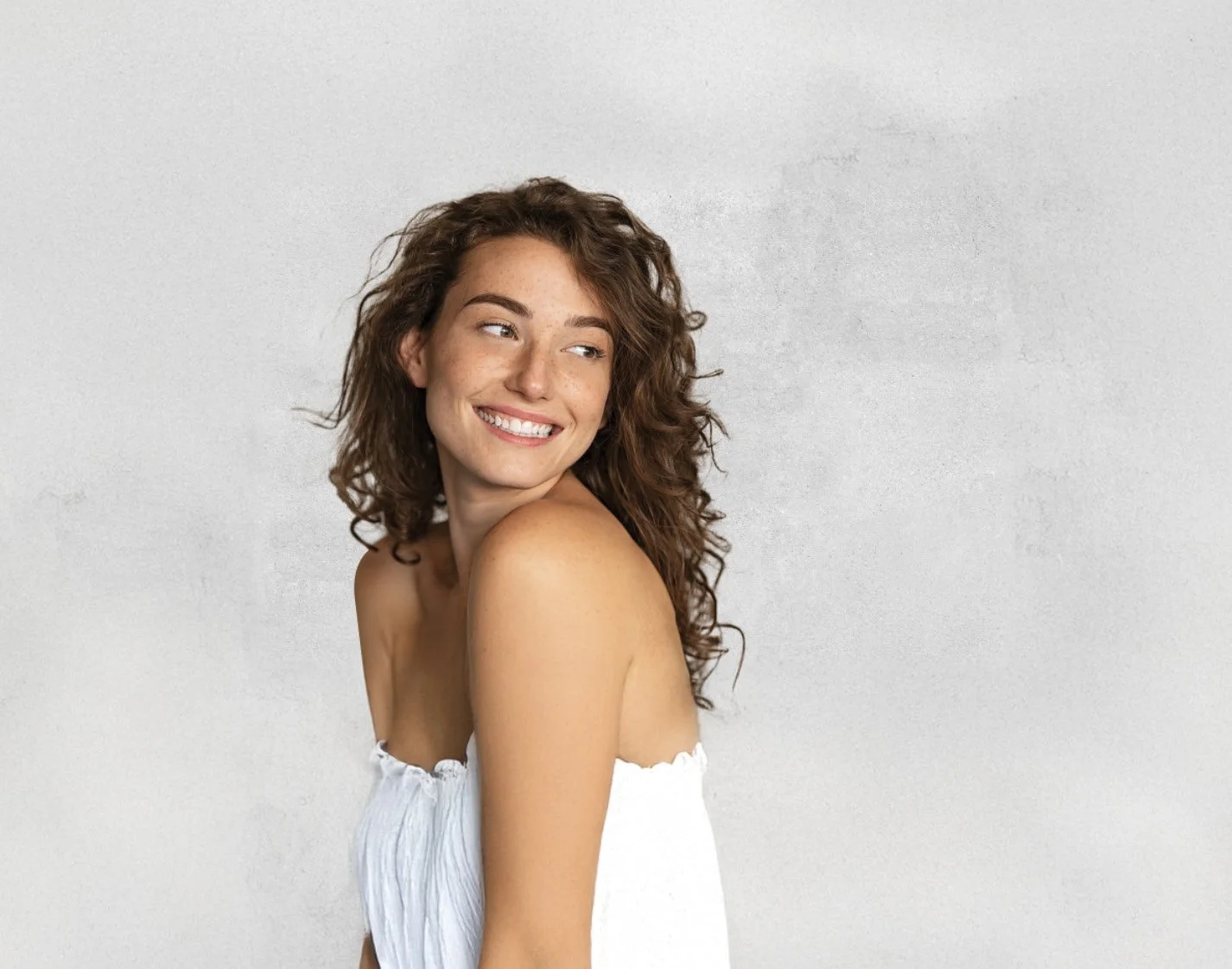 A smiling woman with long curly brown hair looking over her shoulder, wearing a white strapless top against a plain light gray background.