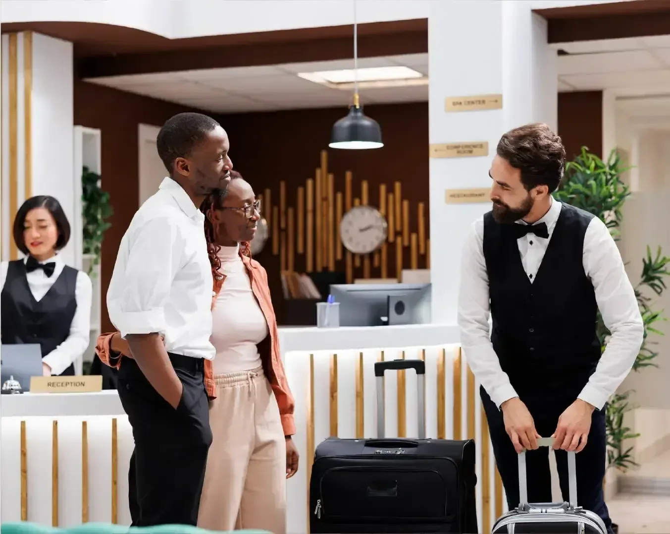 Hotel reception area with a staff member at the counter, four guests with luggage, and plants in the background.
