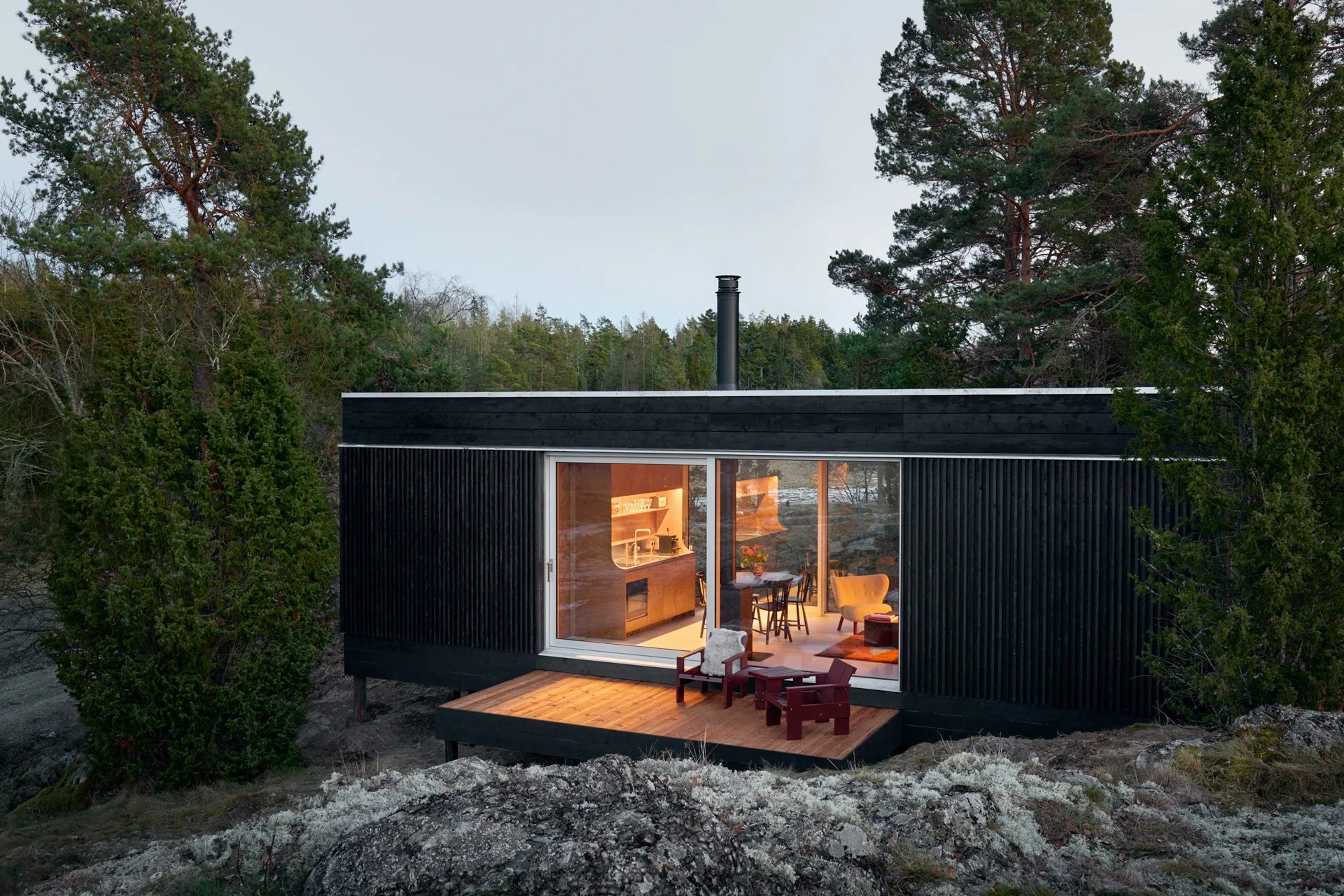 Modern black house on rocky terrain surrounded by trees, with interior lights visible through large glass sliding doors, outdoor deck with chairs.