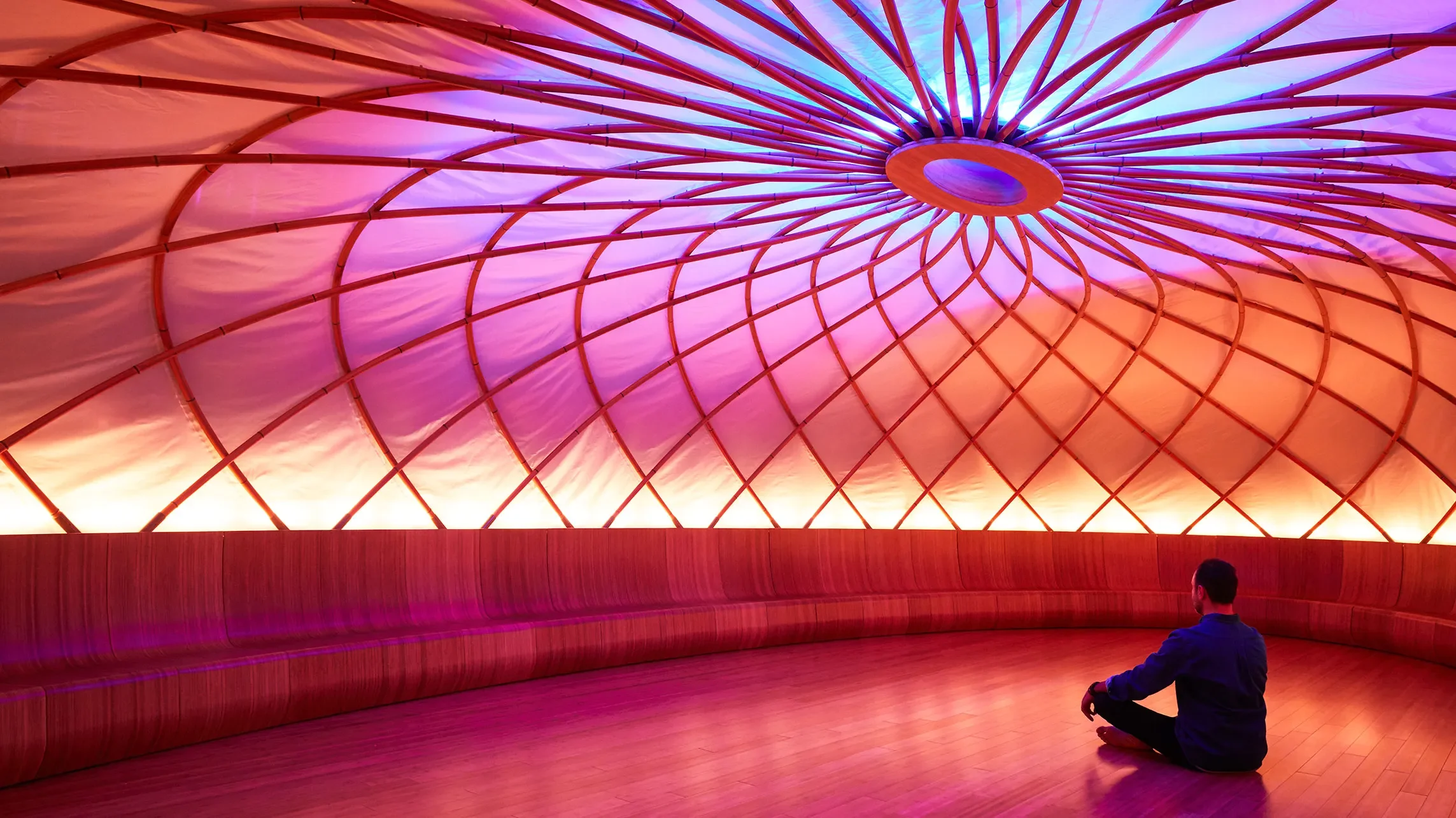 A person sitting cross-legged inside a circular room with a colorful, abstract ceiling and a wooden floor.