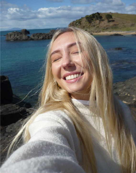 A smiling blonde woman taking a selfie near a rocky shoreline with blue water and a green hill in the background.