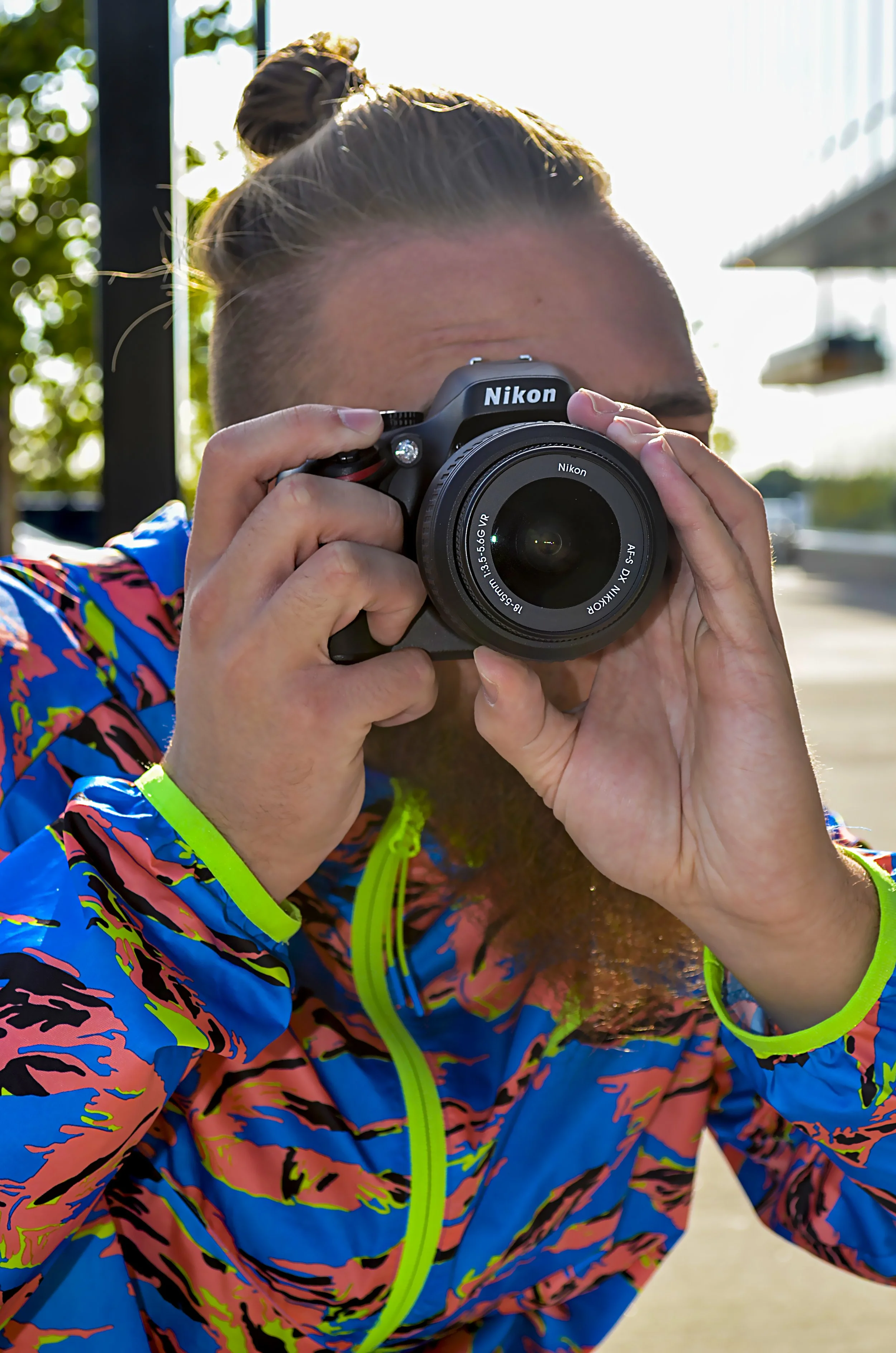 Close-up of a man in a colourful jacket holding a camera and aiming it directly toward the photographer, preparing to take a shot.