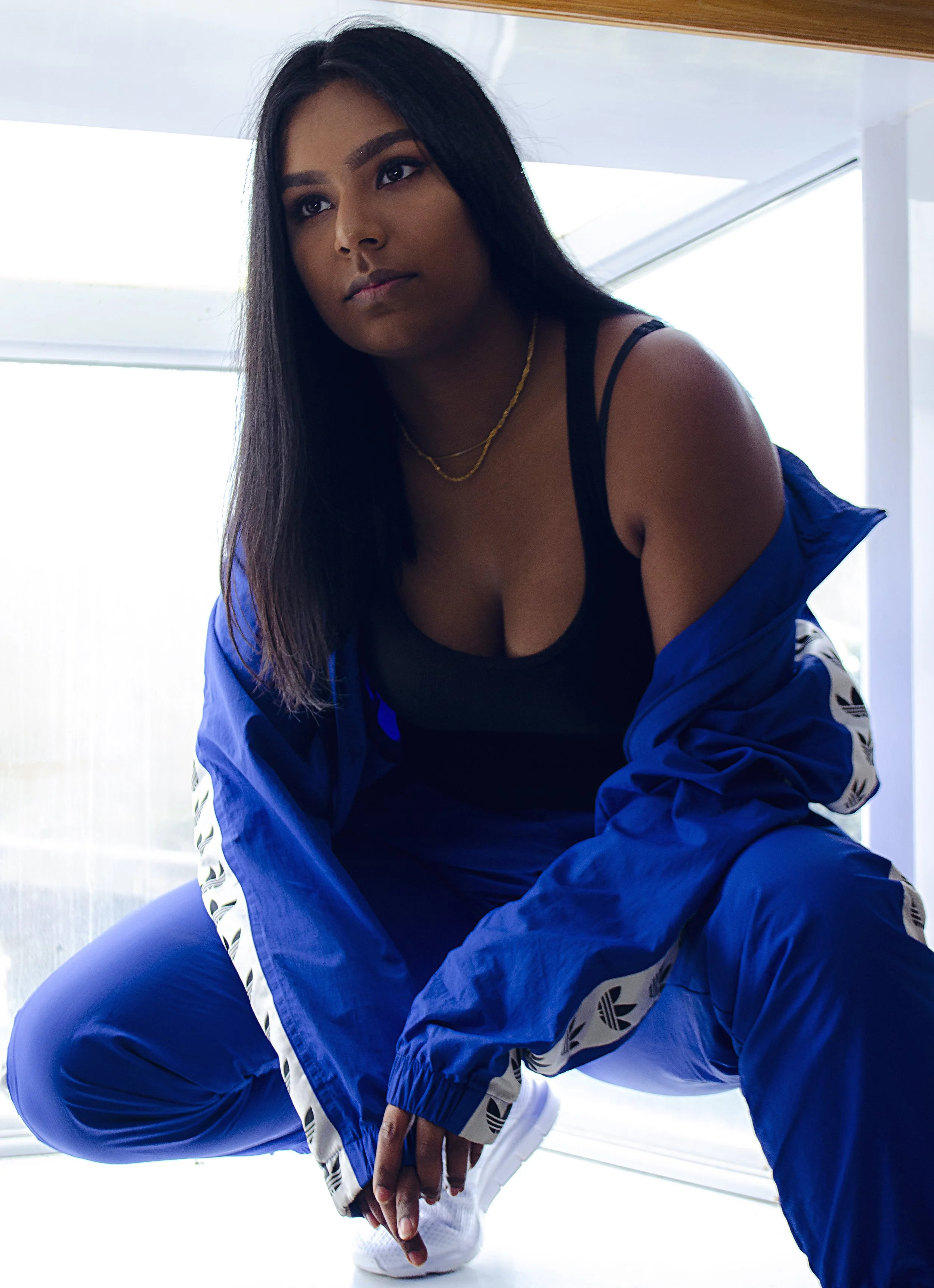 A woman crouches in front of a window as bright white light streams through. She looks off to the side, away from the camera. She wears an all-blue Adidas tracksuit with the sleeves slipped down, revealing one shoulder and a black tank top underneath