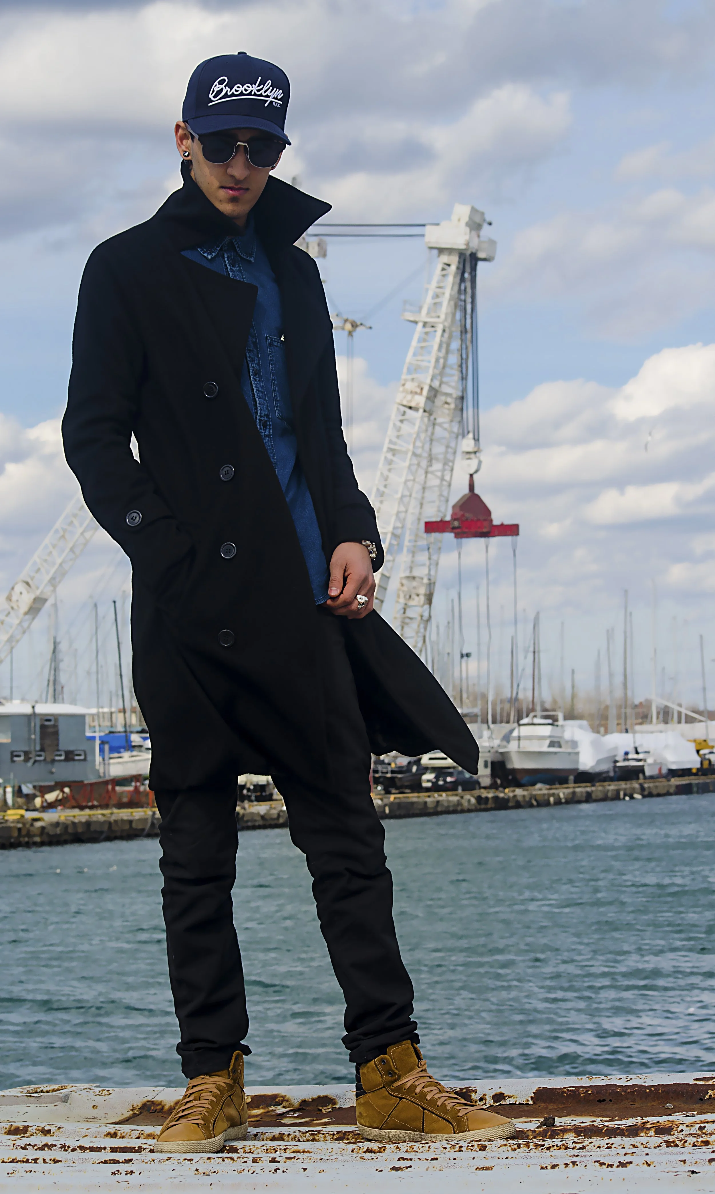 A man stands on a rusted structure facing the camera, with a body of water behind him. He is wearing a baseball cap, glasses, and a long black overcoat that billows in the wind.