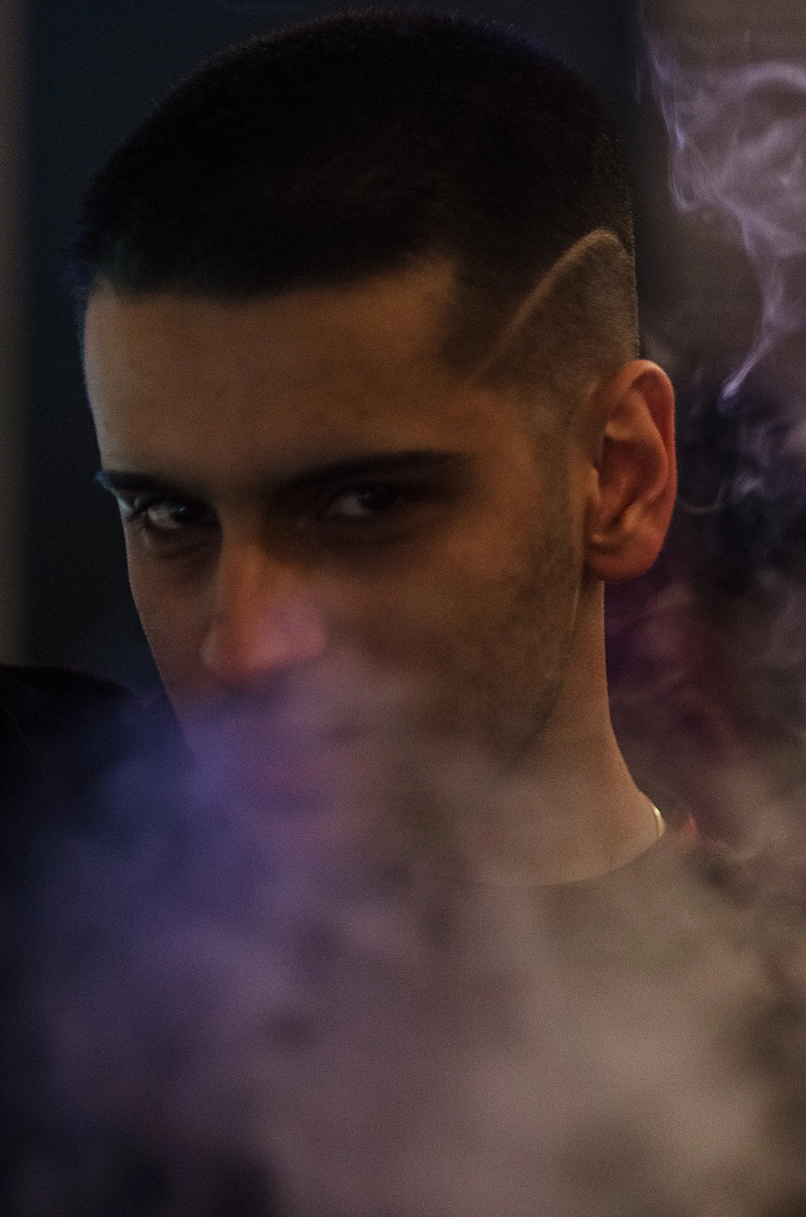 A close-up portrait of a man looking directly into the camera. Exhaled smoke partially obscures his face, while blue and purple lighting casts dramatic shadows and highlights across the scene.