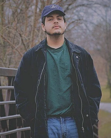 A man stands on a bridge facing the camera in a three-quarter portrait pose with his hands in his pockets. He is wearing a blue baseball cap, blue jeans, a loose-fitting black coat worn unzipped, and a green shirt underneath.