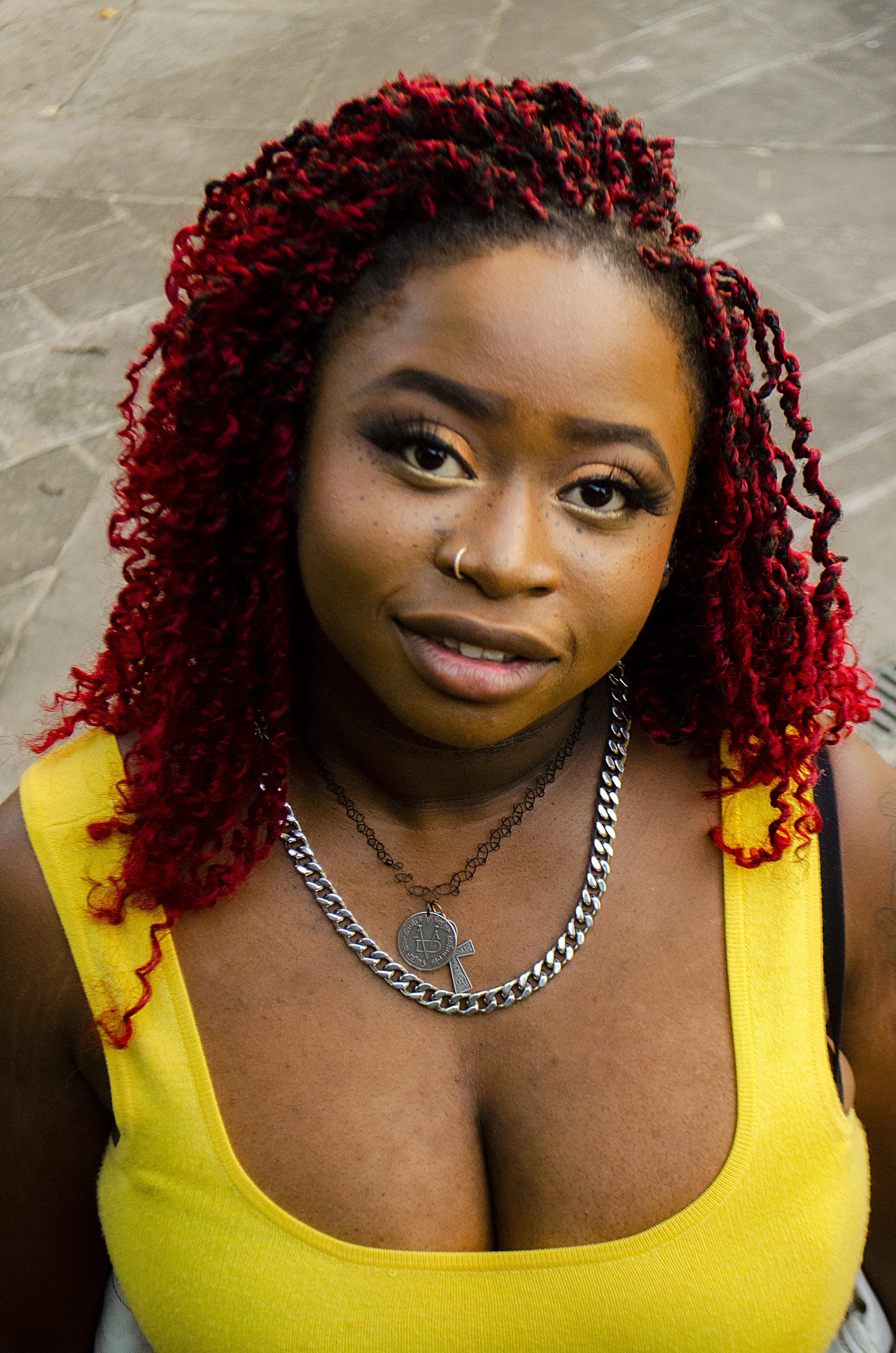 A woman is photographed from a slightly elevated angle in a close-up portrait. She looks up toward the camera with her head tilted, wearing a yellow tank top, layered silver necklaces, and her red curly hair framing her face.