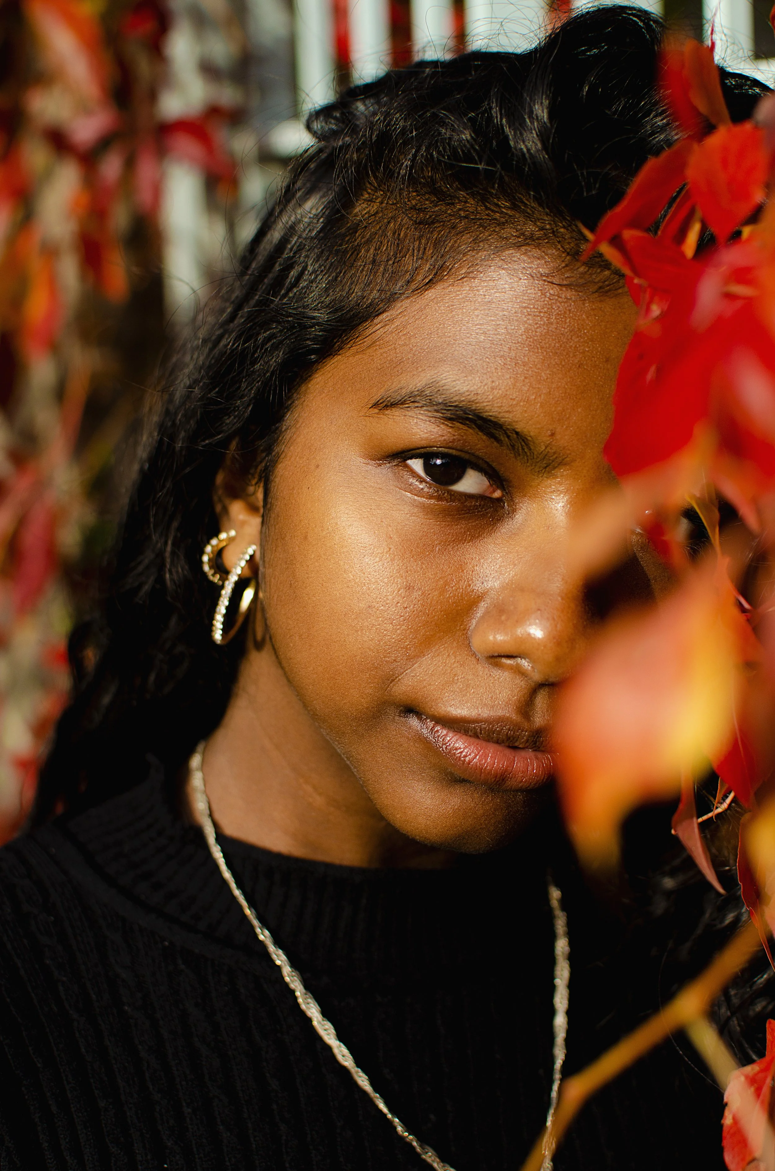 A close-up headshot of a woman looking toward the camera with her head slightly tilted. Warm red autumn leaves partially cover one eye while the other looks directly at the viewer. She wears all black clothing, with flowing black hair, a thin gold ch