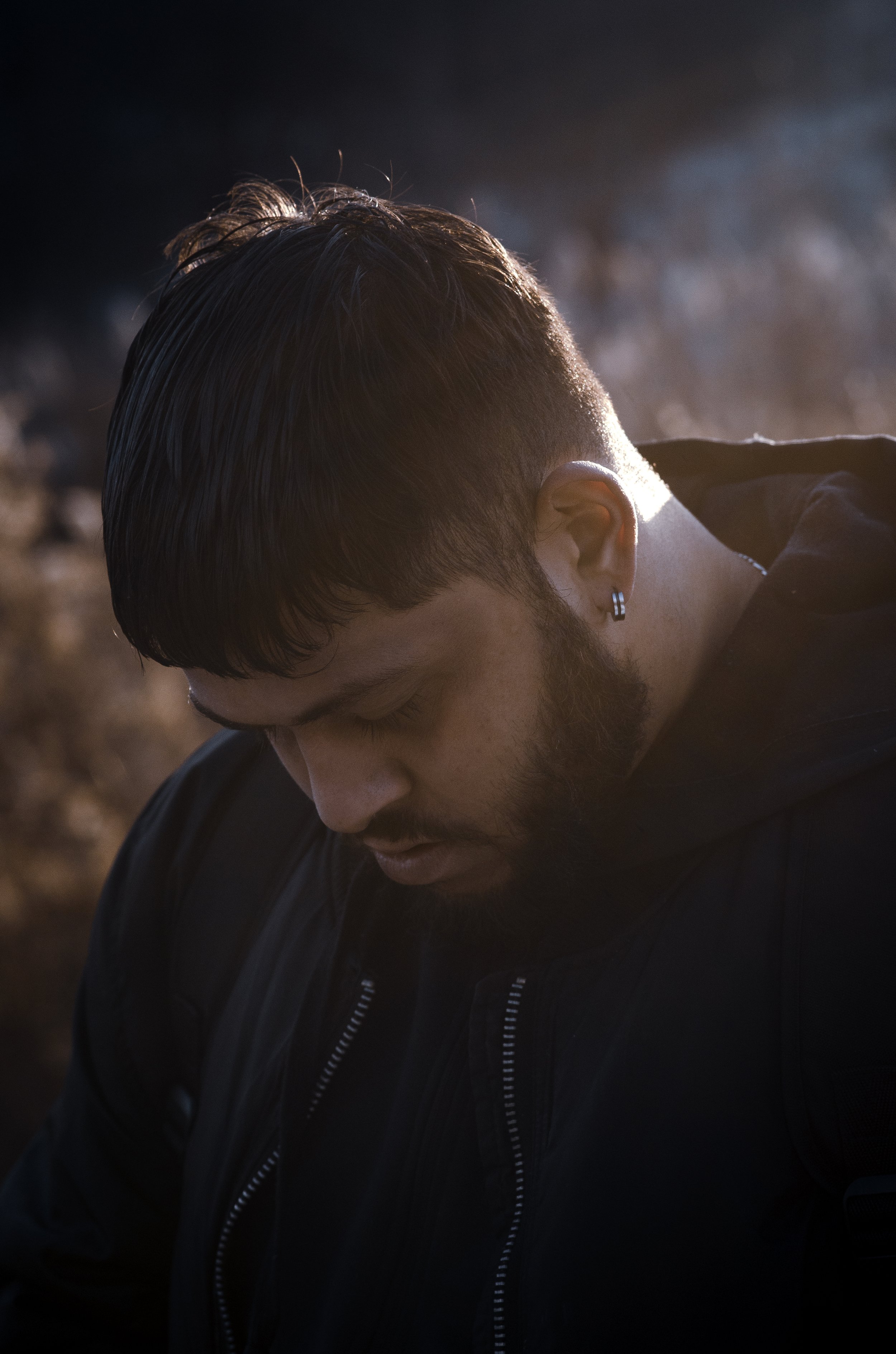 A headshot portrait of a man looking downward, dressed in black clothing. Soft morning sunlight filters through a light mist surrounding him, creating a gentle and atmospheric scene.
